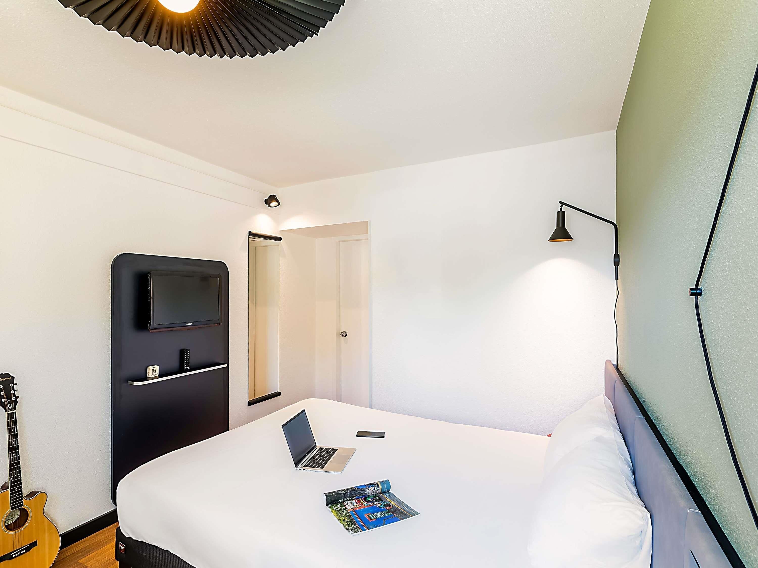 A hotel room with a bed, a desk, a laptop, a guitar, and a TV on a stand.