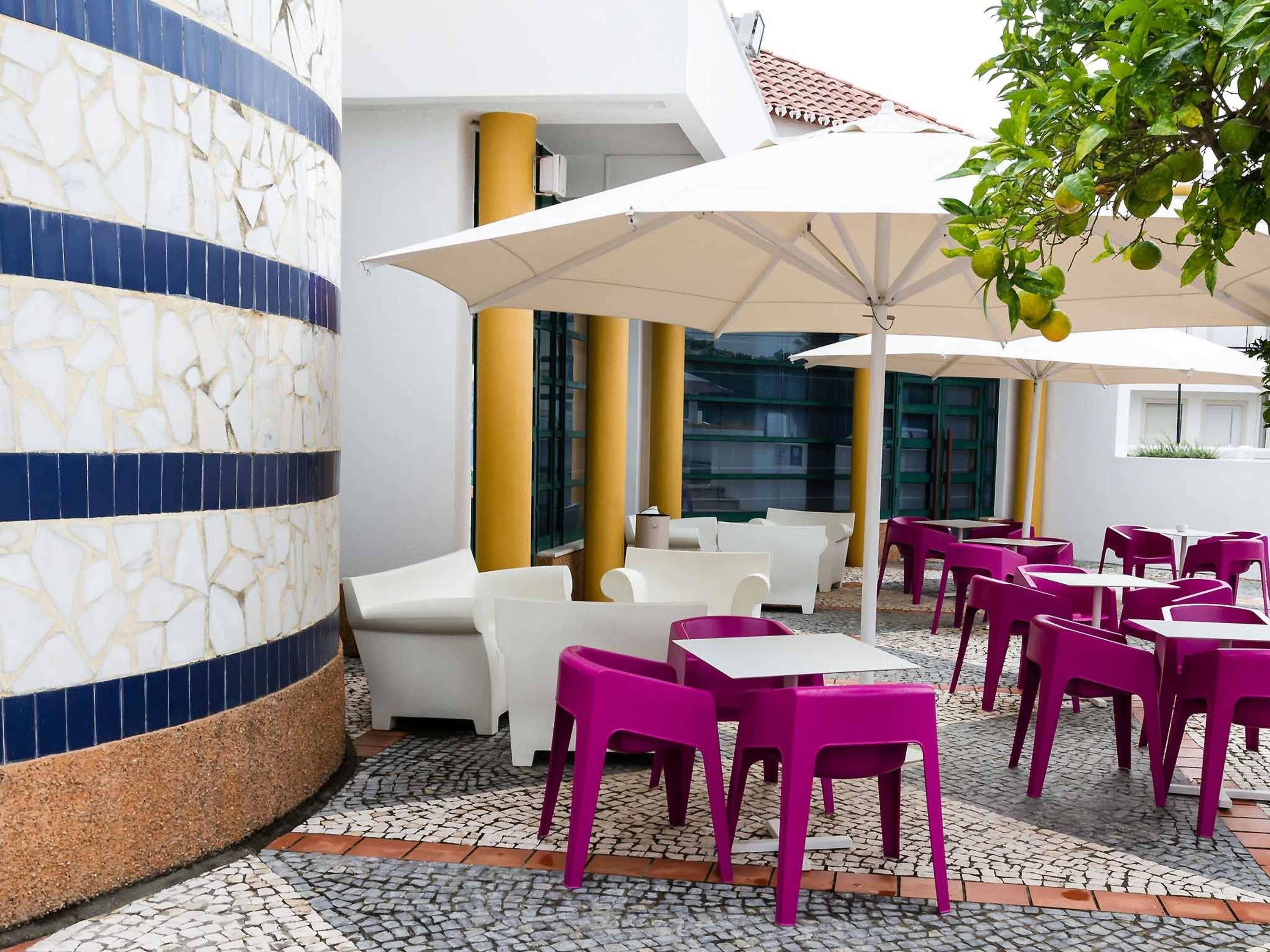 An outdoor seating area with white and pink chairs, large umbrellas, and a tiled floor.