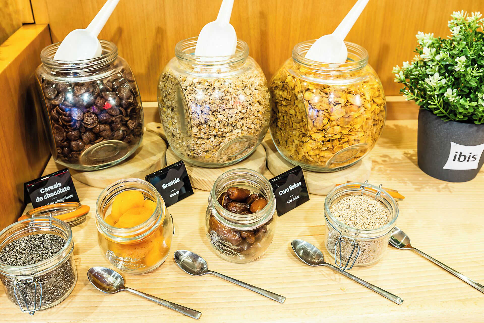 A variety of food items in jars with labels, including cereals, granola, and corn flakes, displayed on a wooden surface.