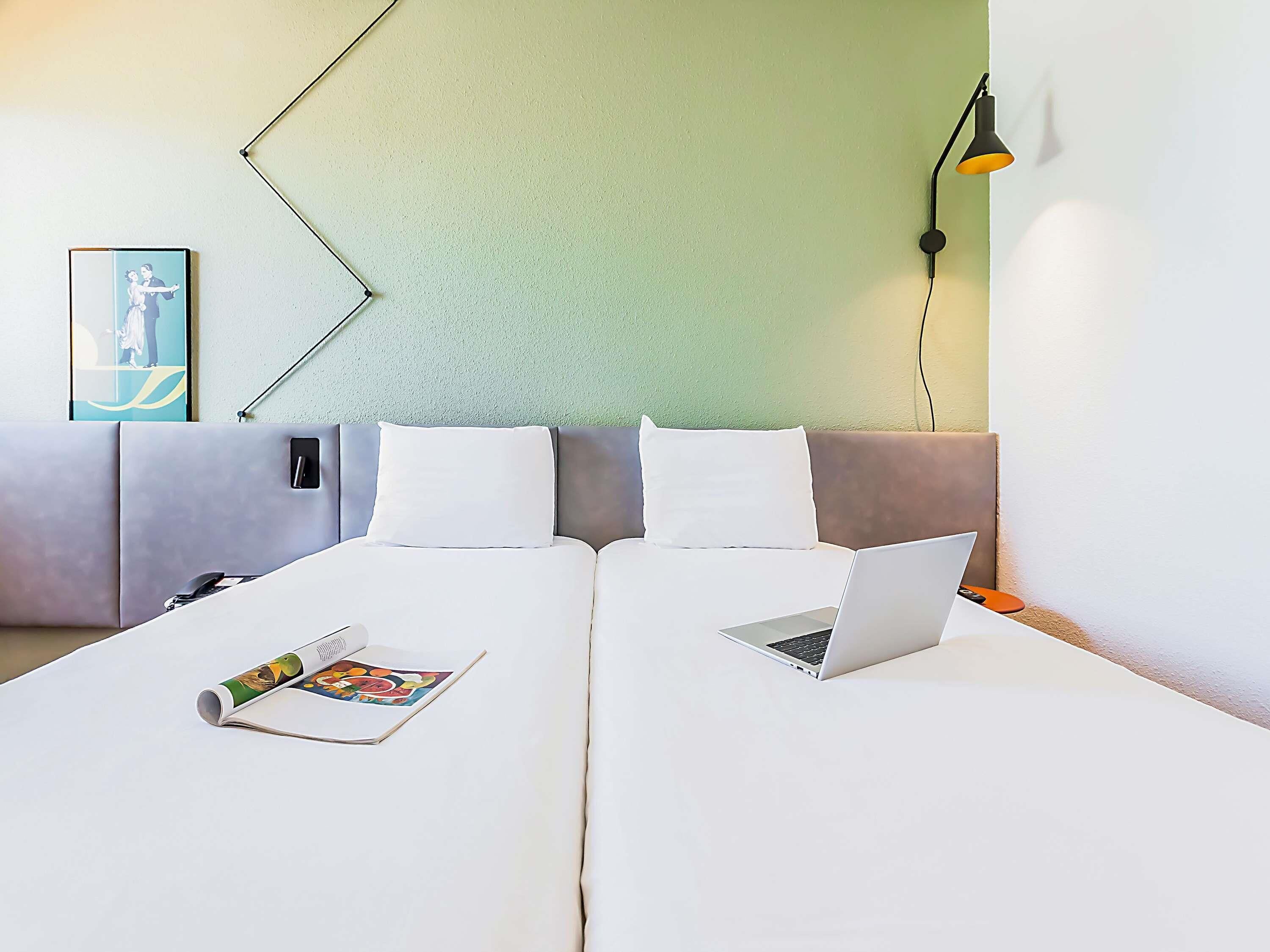 A double bed with a laptop and a book on it, a bedside table with a lamp, and a wall with a picture frame.
