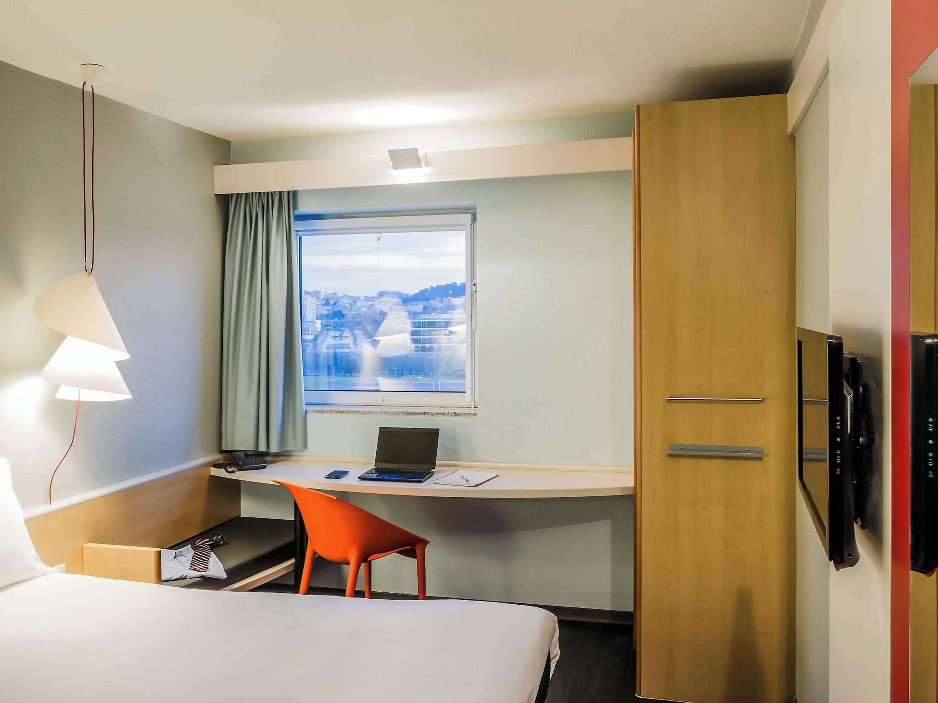 Habitación de hotel con cama, escritorio con ordenador portátil, silla, ventana con vista a la ciudad y armario.