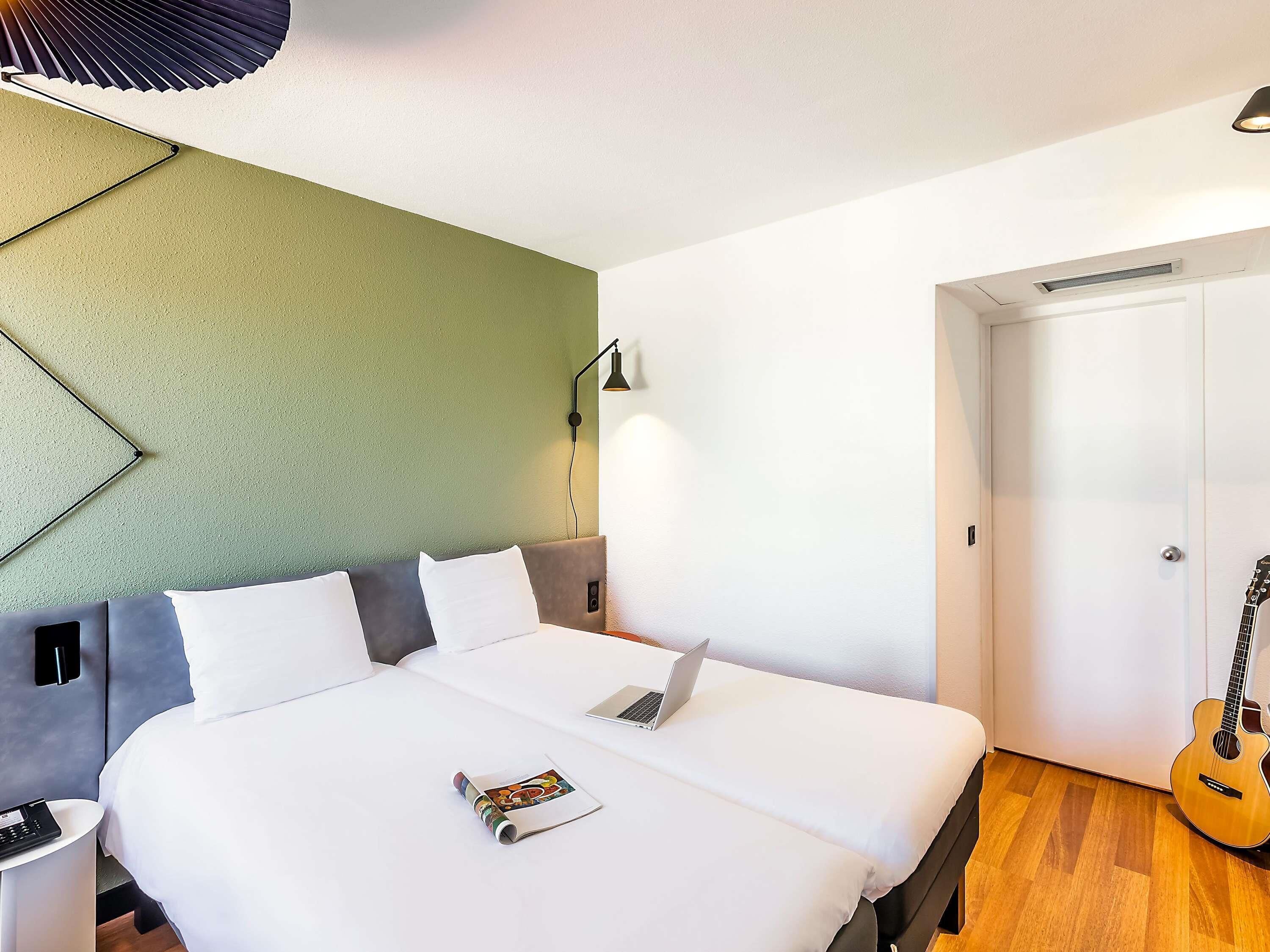 A hotel room with a bed, a laptop, a guitar, and a wall-mounted lamp.