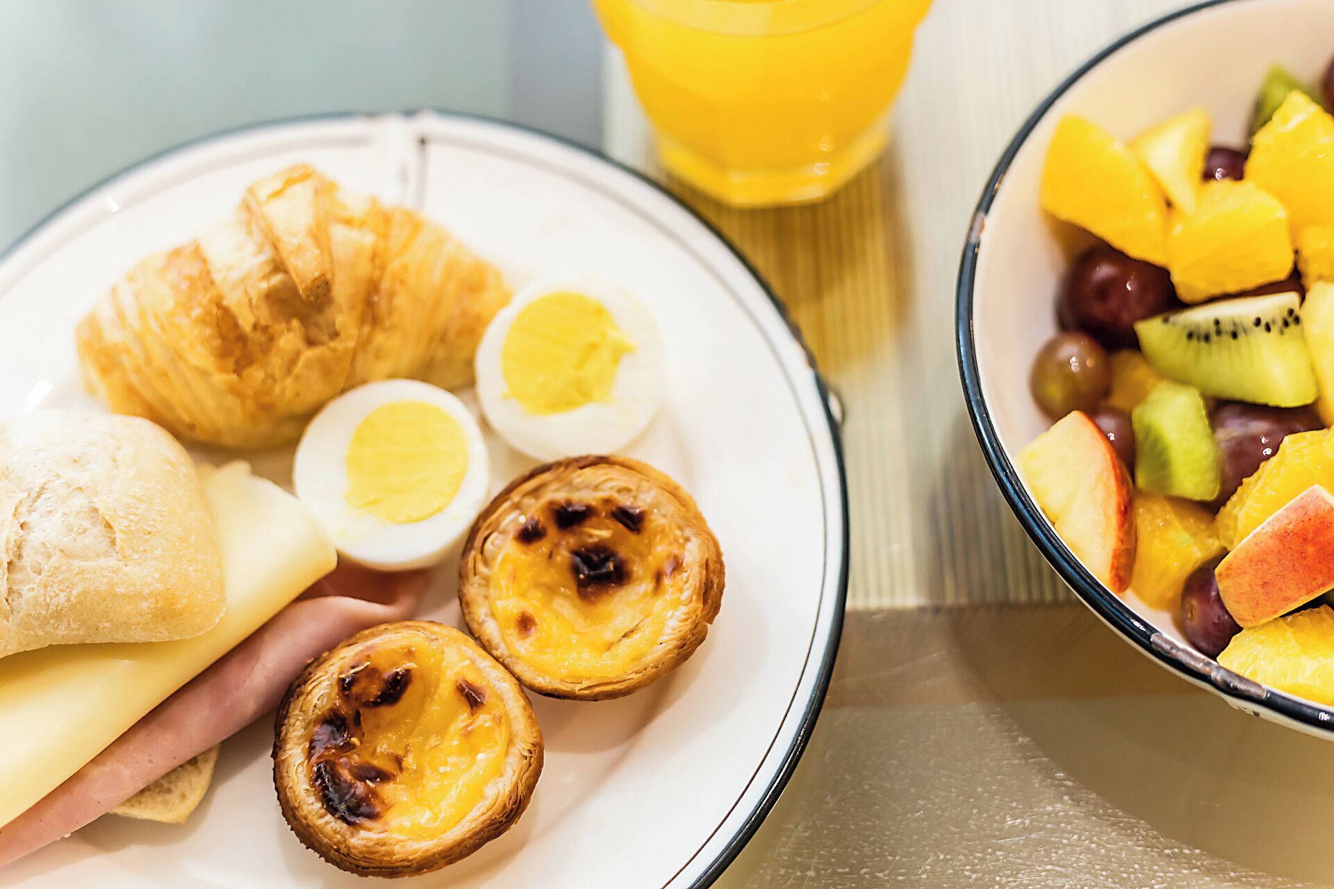 Een ontbijt bord met croissants, ham, kaas en eiertaartjes, en een kom fruit.