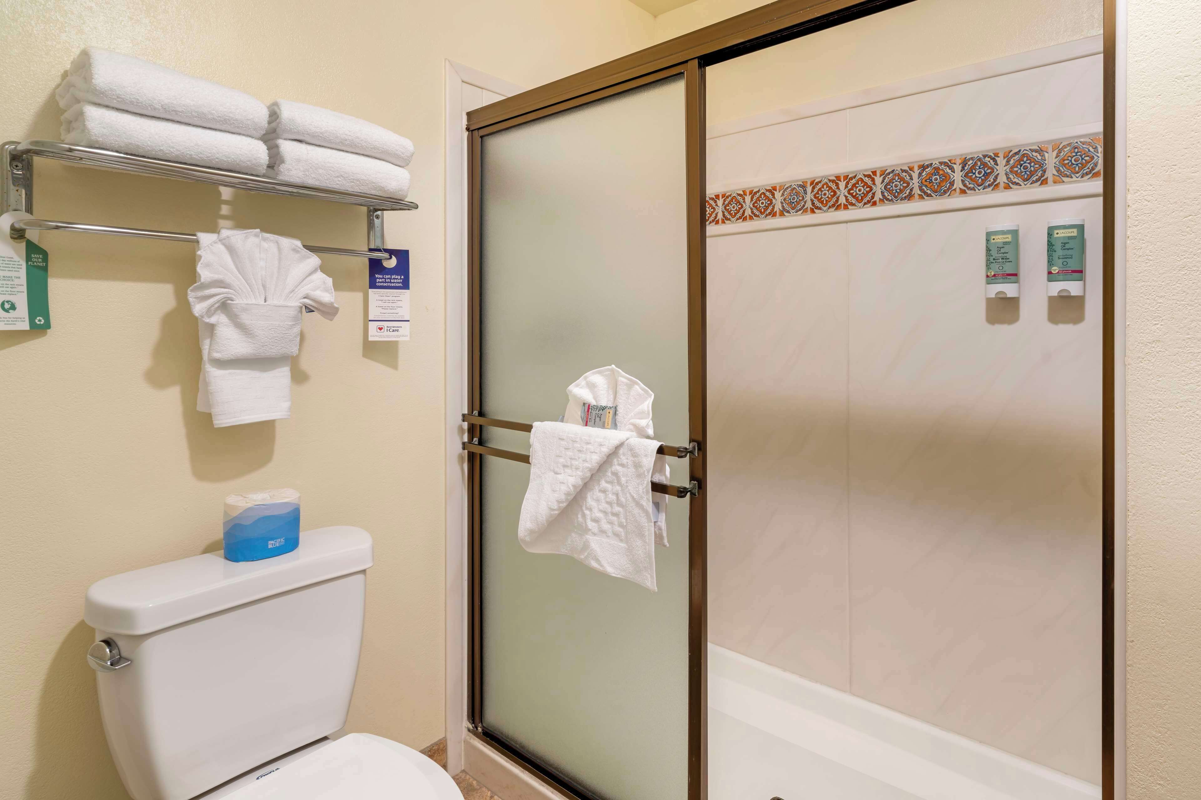A bathroom with a toilet, a towel rack with folded towels, a shower with a glass door, and a toilet paper dispenser.