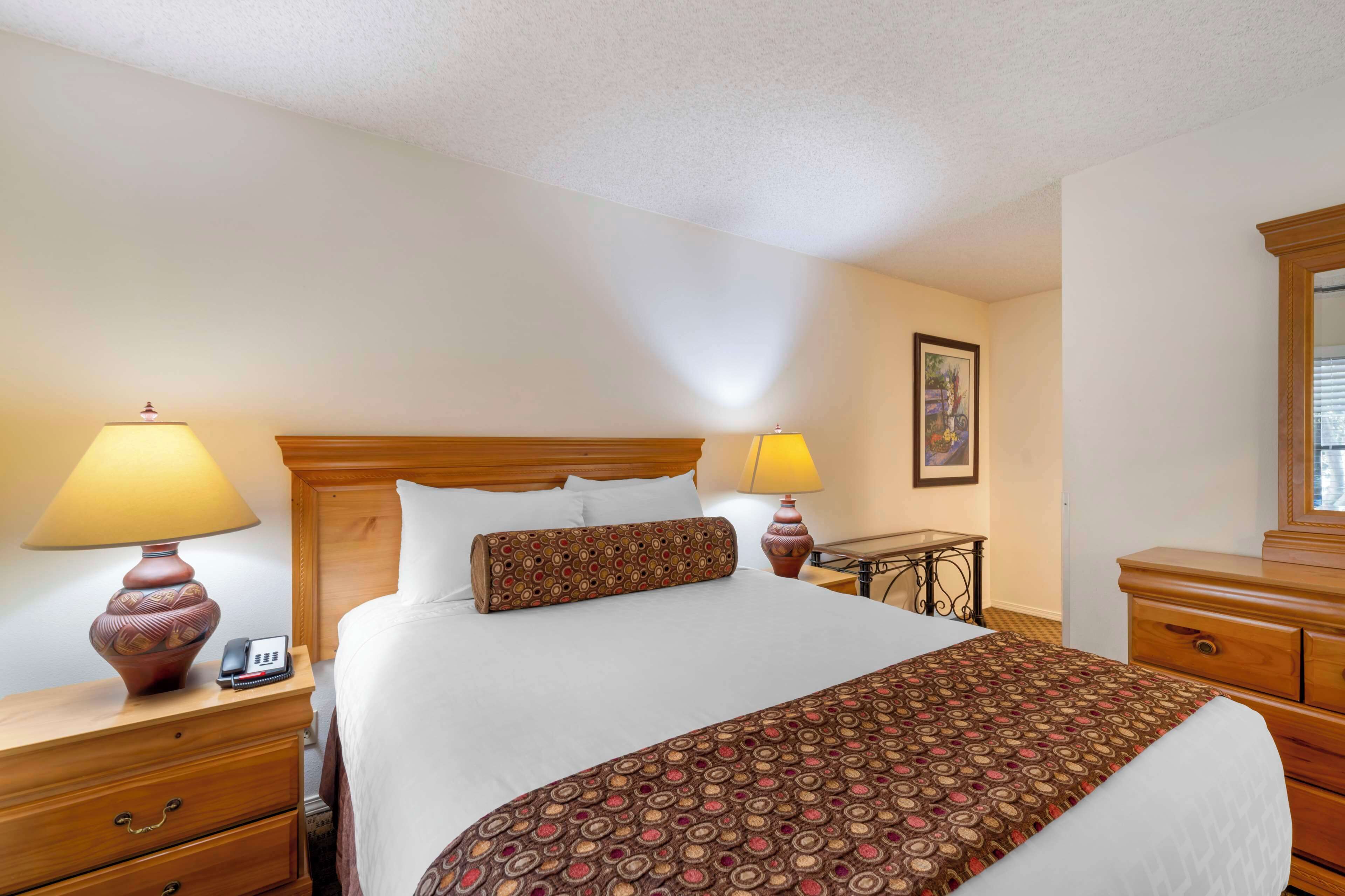 A hotel room with a large bed, two bedside lamps, a wooden nightstand, a small table, and a dresser.