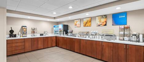 Uma área de pequeno-almoço self-service com diversas opções de comida, uma máquina de café e um frigorífico.