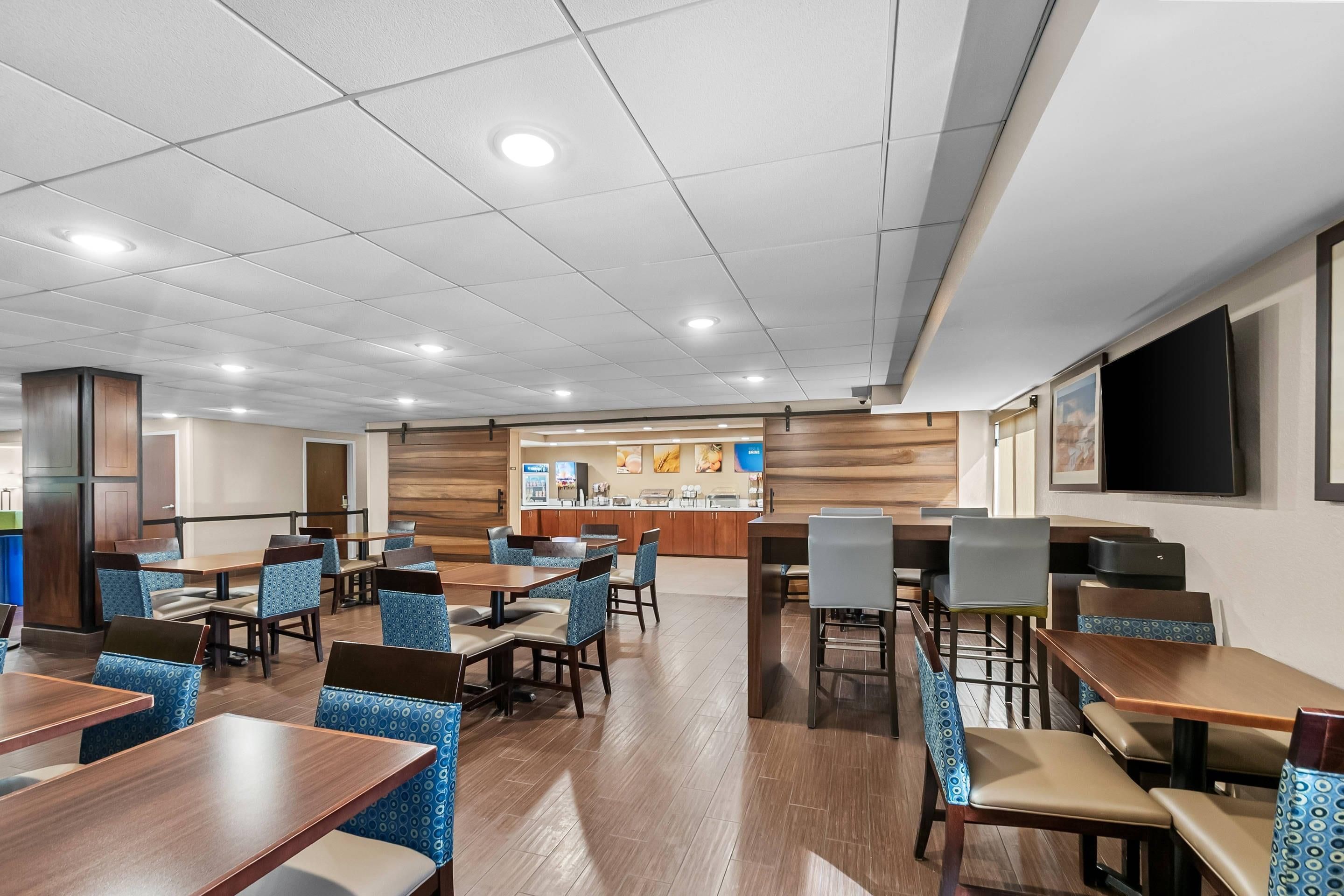 A hotel dining area with multiple tables and chairs, a counter with a menu display, and a television screen mounted on the wall.