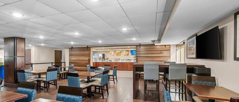 A hotel dining area with multiple tables and chairs, a counter with a menu display, and a television screen mounted on the wall.