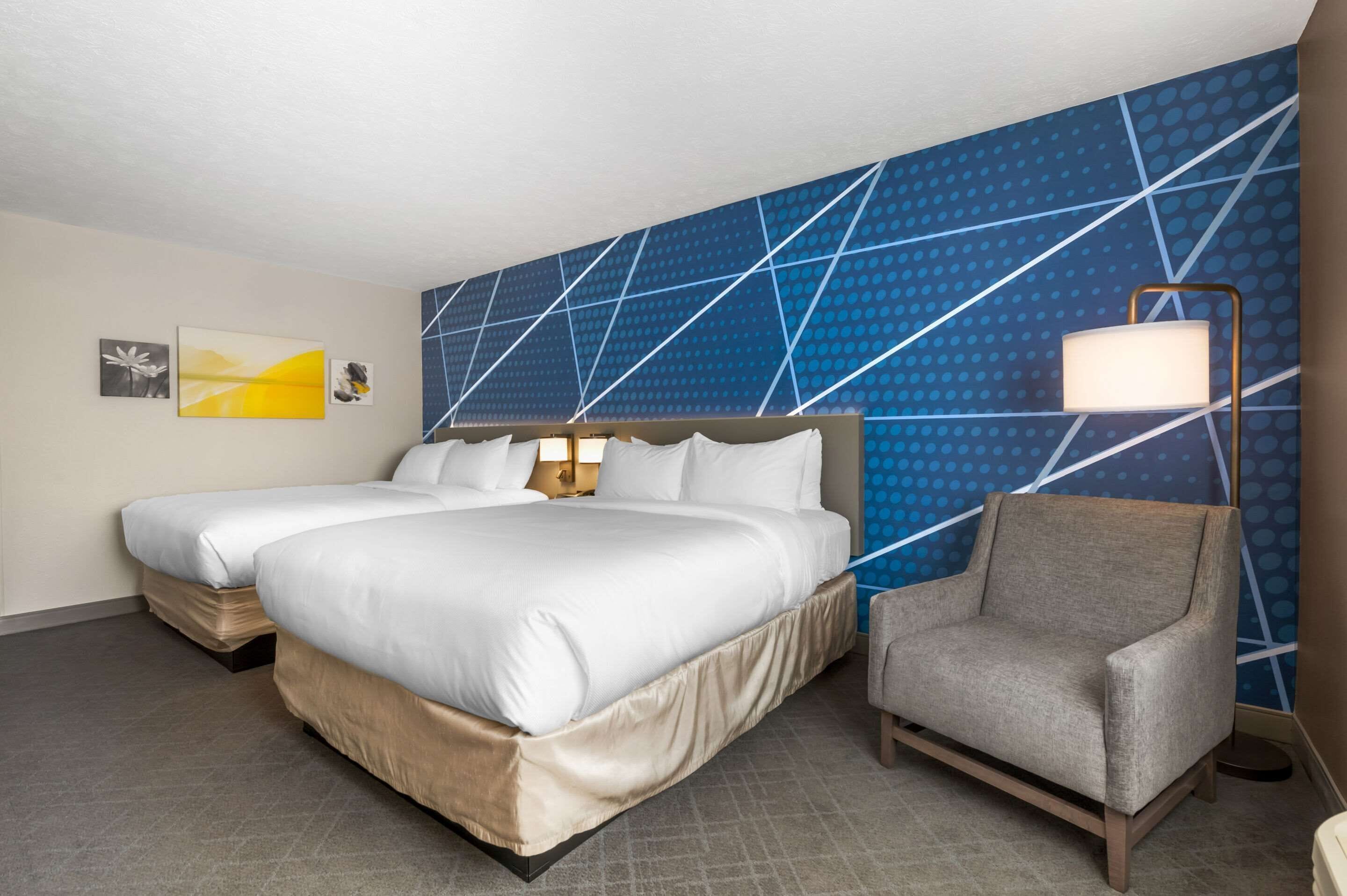 A hotel room with two beds, a nightstand, a lamp, and a chair.