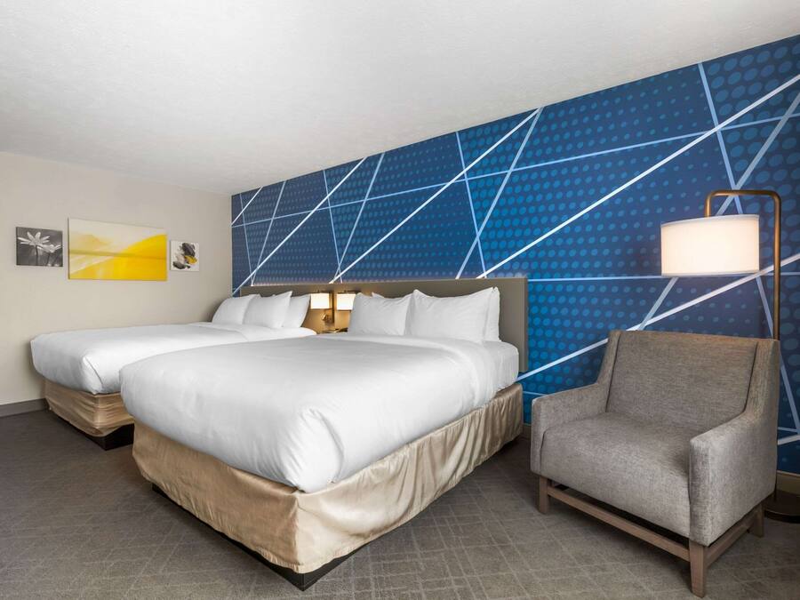 A hotel room with two beds, a nightstand, a lamp, and a chair.