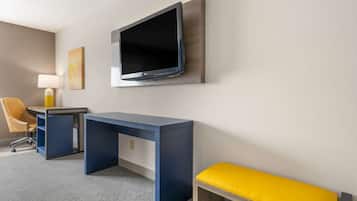 A hotel room with a desk, chair, TV, lamp, and a yellow bench.