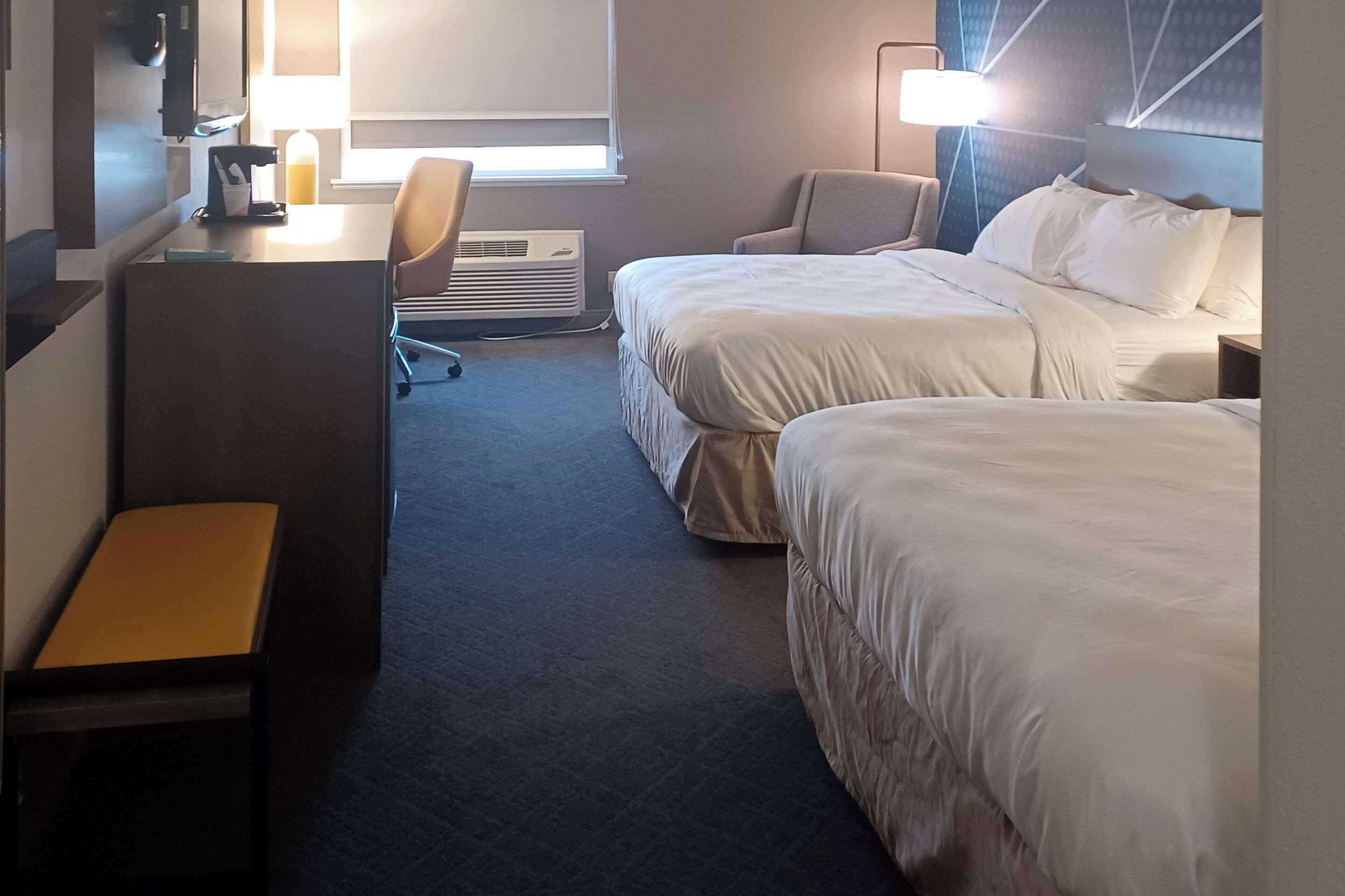 Habitación de hotel con dos camas, un escritorio con silla, una cafetera y una ventana con persianas.