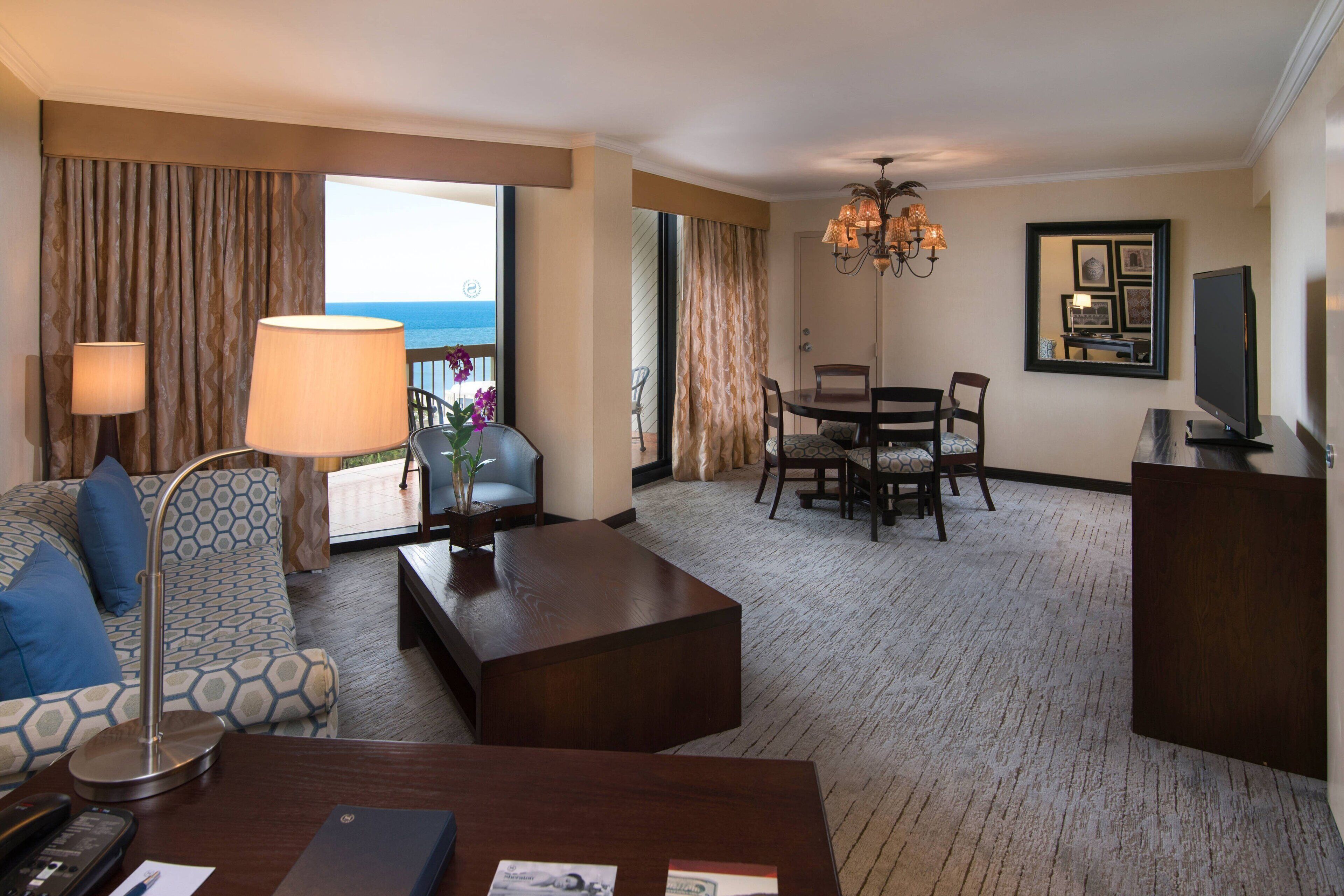 A hotel room with a sofa, coffee table, dining table, chairs, a TV, and a large window with curtains.