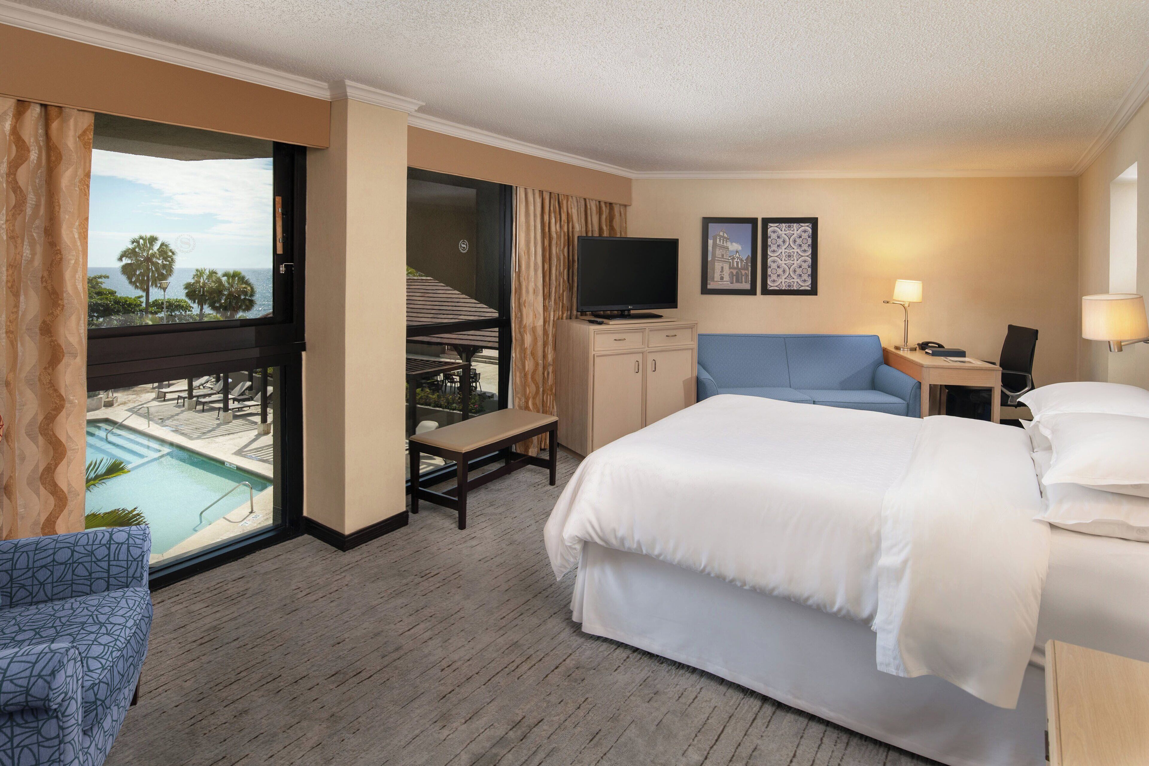 Habitación de hotel con una cama grande, un escritorio con computadora, televisión, balcón con vista a la piscina y una silla.
