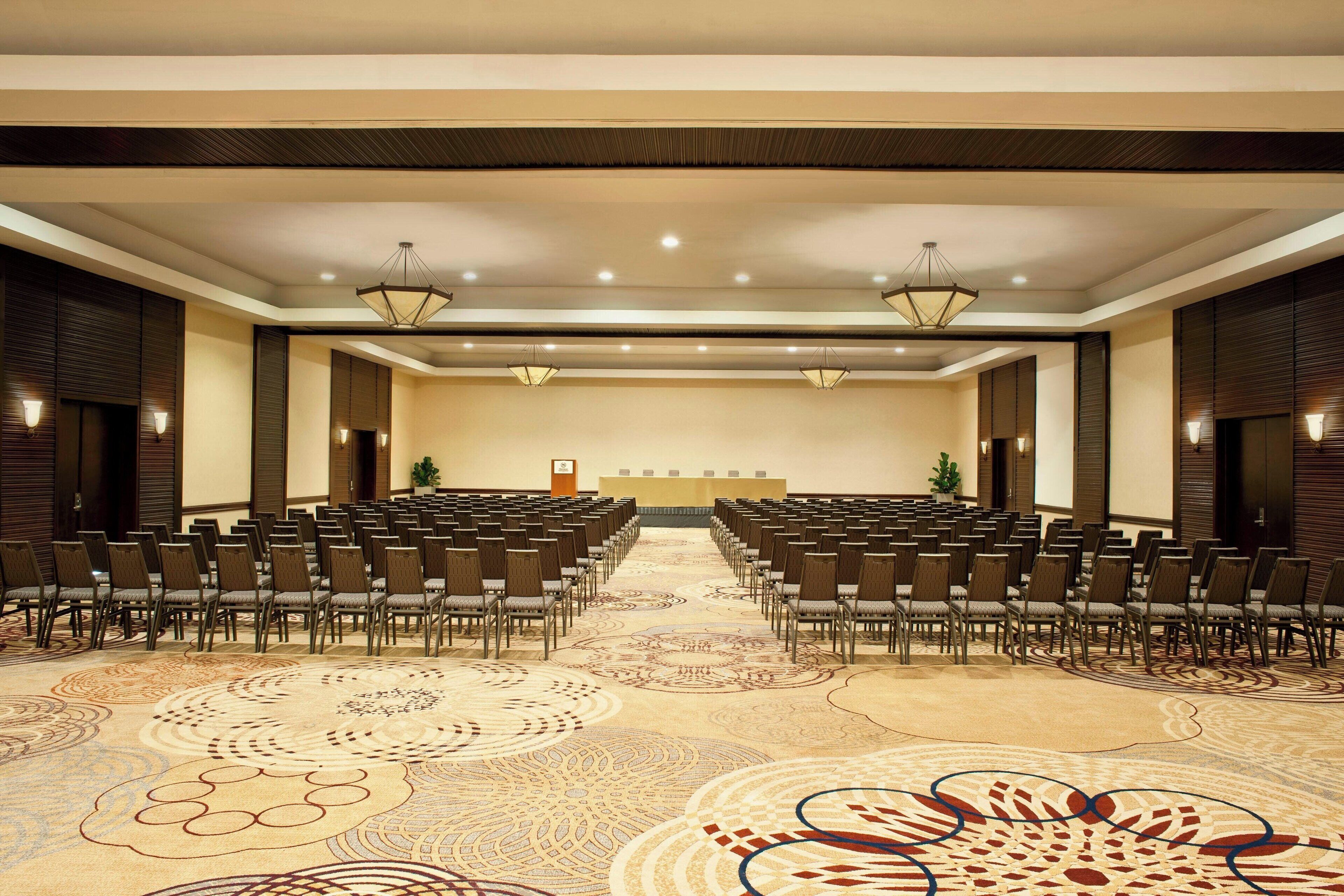 A conference room with rows of chairs facing a stage, a patterned carpet, and wall-mounted lighting.