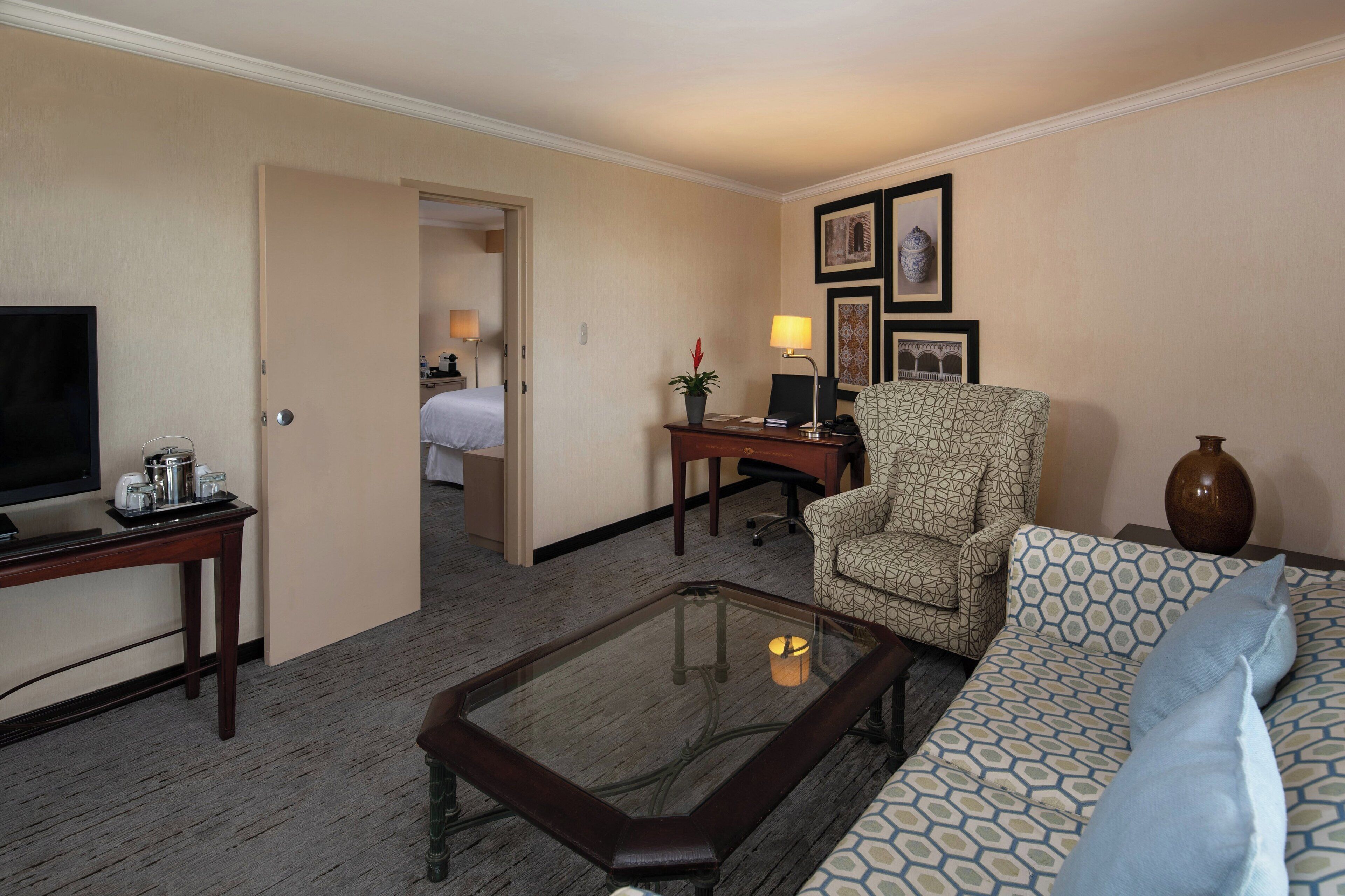 A hotel room with a glass-top coffee table, a patterned sofa, a patterned armchair, a desk with a lamp, and a television on a stand.