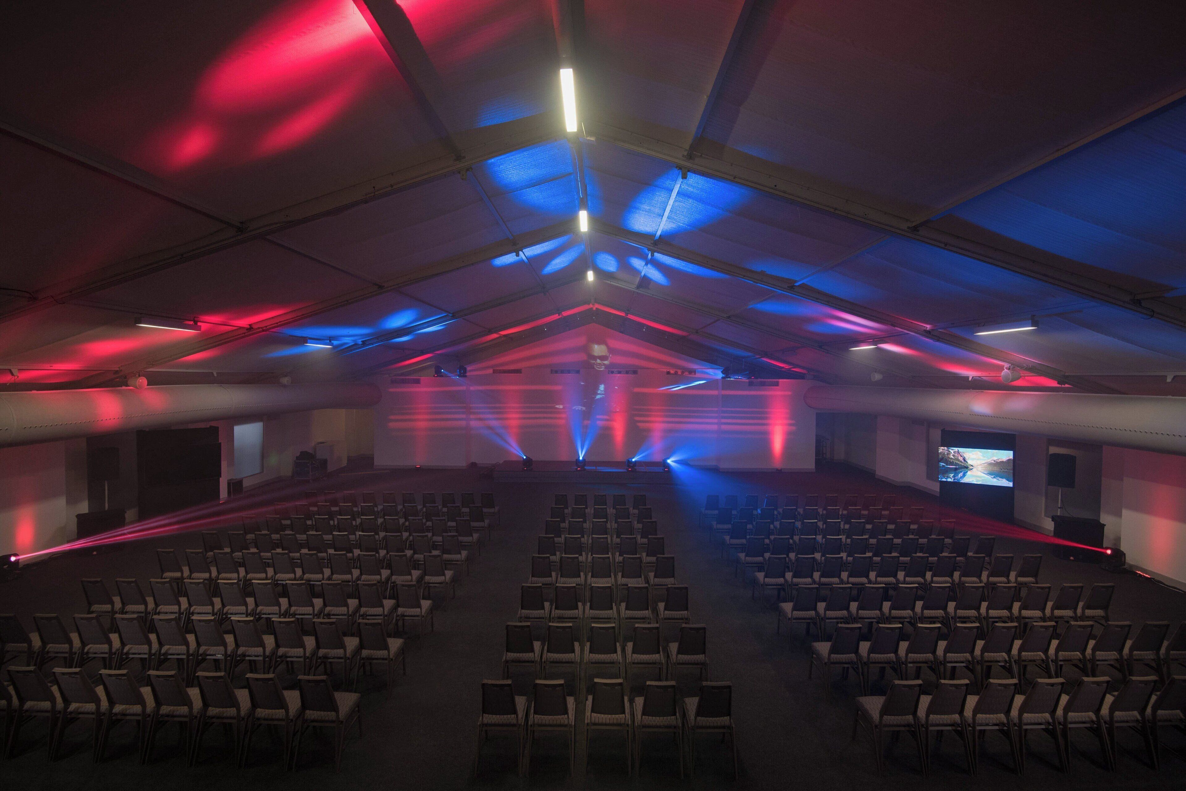 Et indendørs auditorium med rækker af stole, der vender mod en scene, udstyret med et stort skærm og en talerstol.