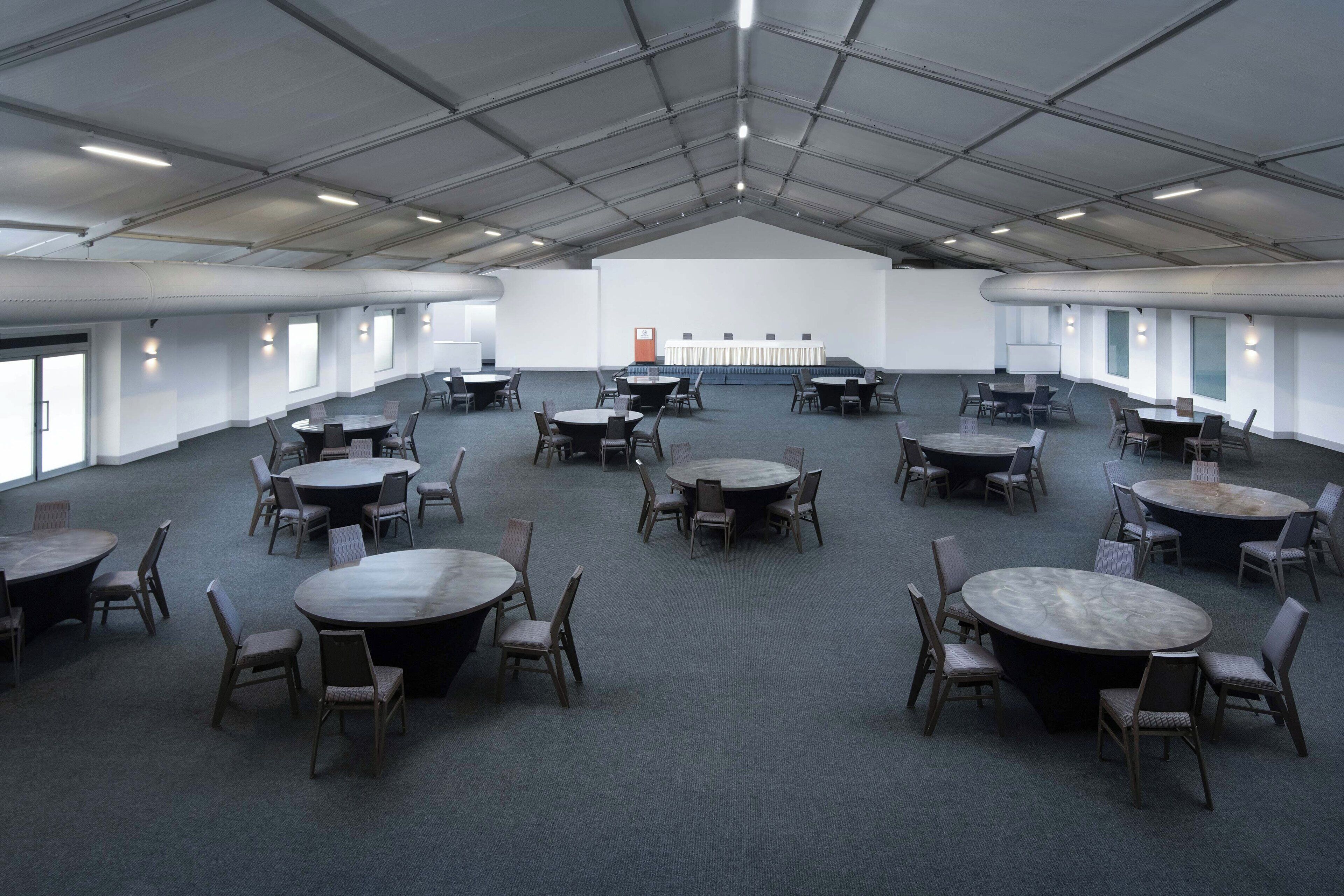 Une grande salle de conférence dotée de nombreuses tables rondes et de chaises, d’une scène avec un pupitre et de grandes fenêtres le long d’un des murs.
