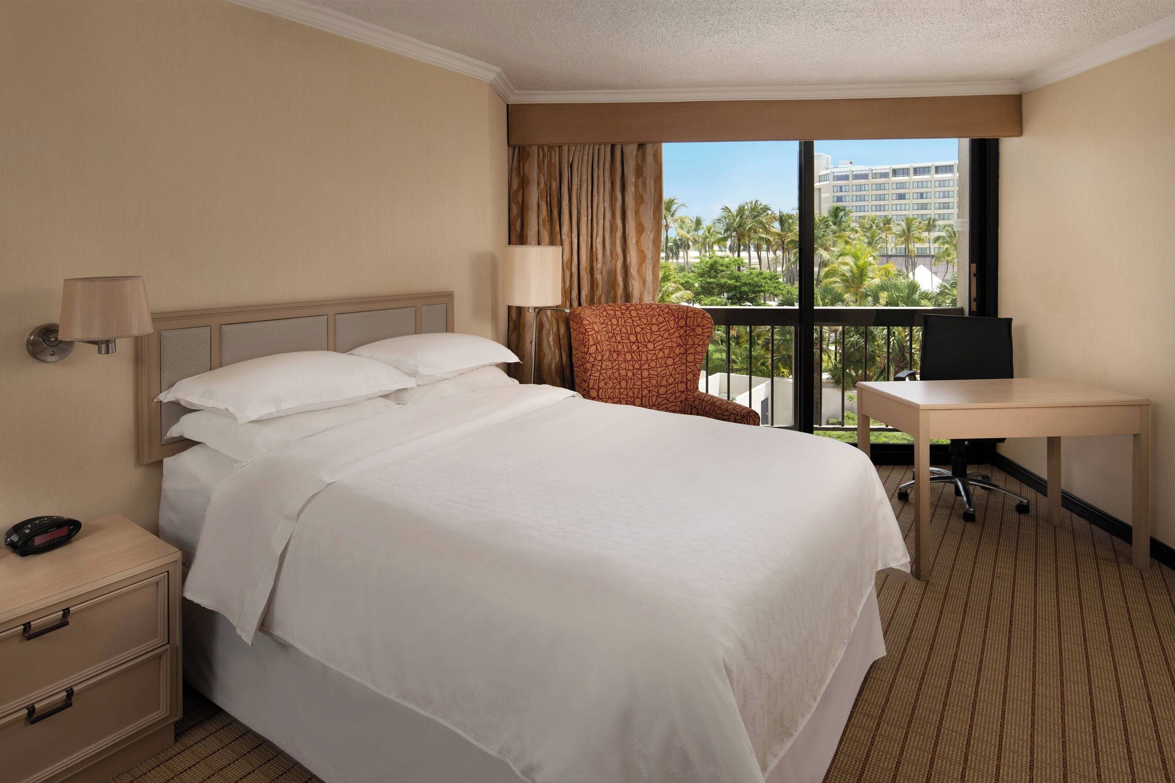 A hotel room with a large bed, a desk, a chair, and a view of palm trees and buildings.