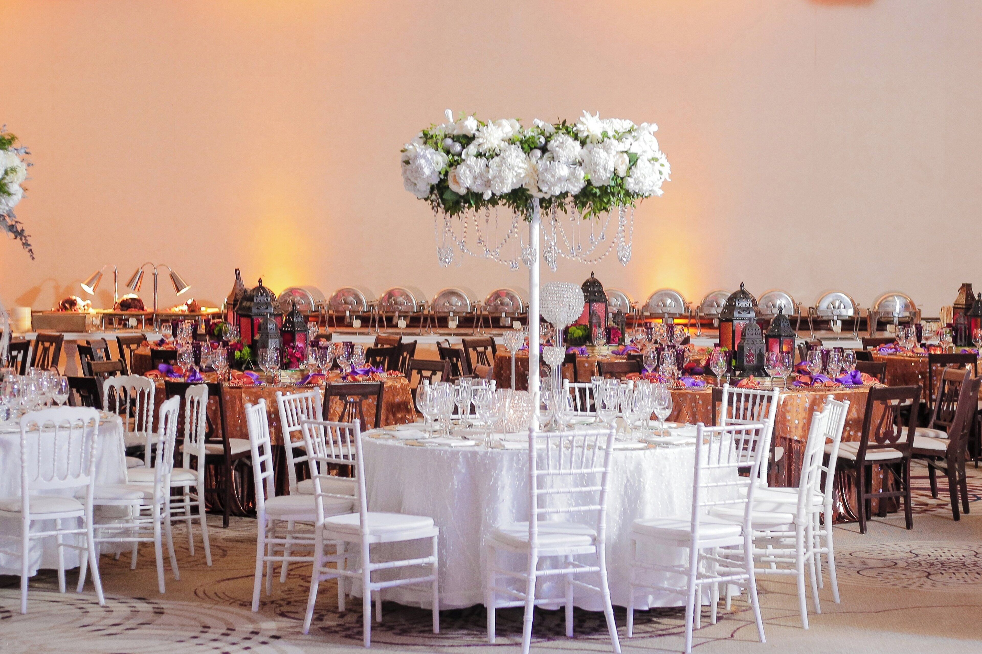 Un salón de banquetes con mesas redondas dispuestas para un evento formal, con sillas y mesas blancas, y un arreglo floral central.