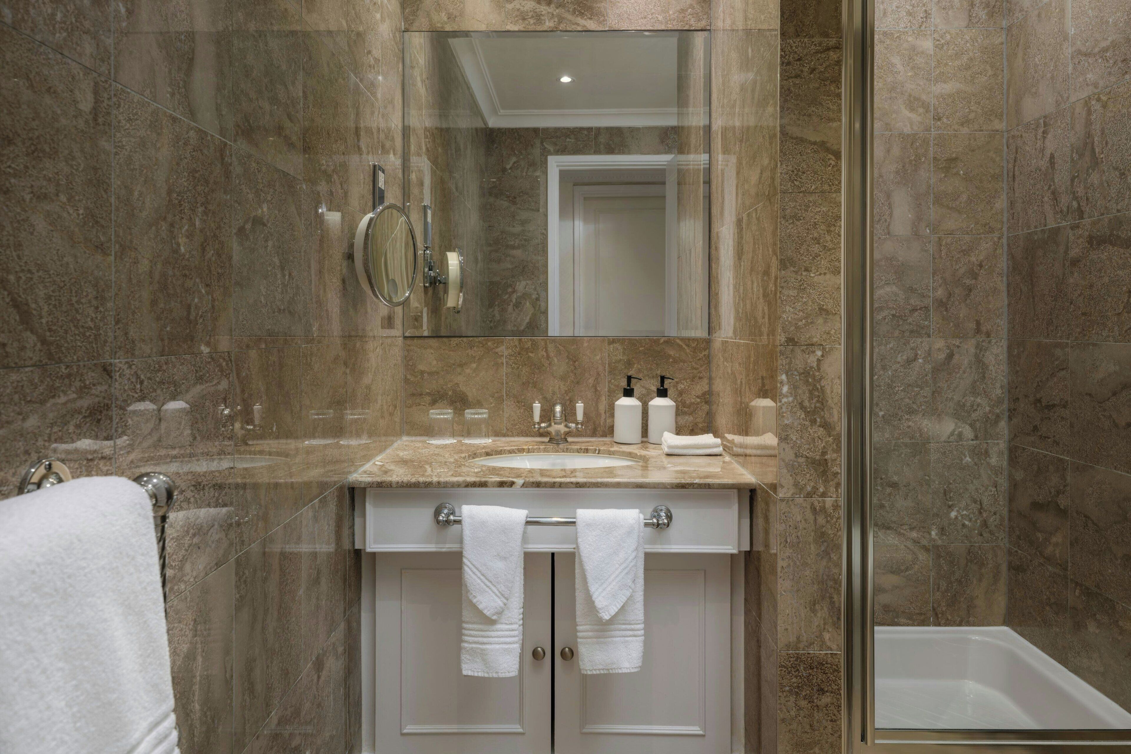 A bathroom with a glass shower enclosure, a sink with toiletries, and towels hanging on the door.