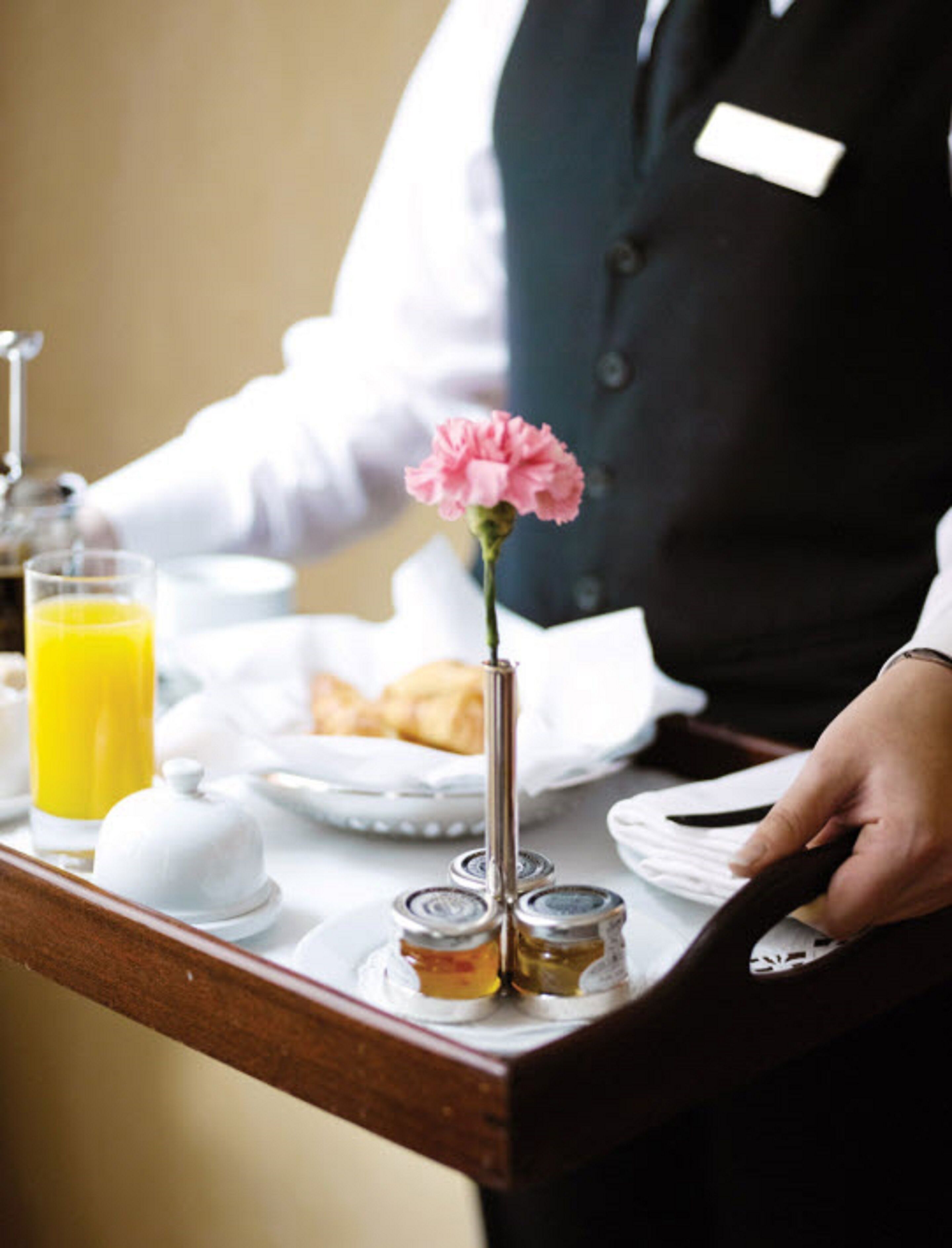 A breakfast tray with a glass of orange juice, a croissant, a small vase with a pink flower, and two jars of jam.