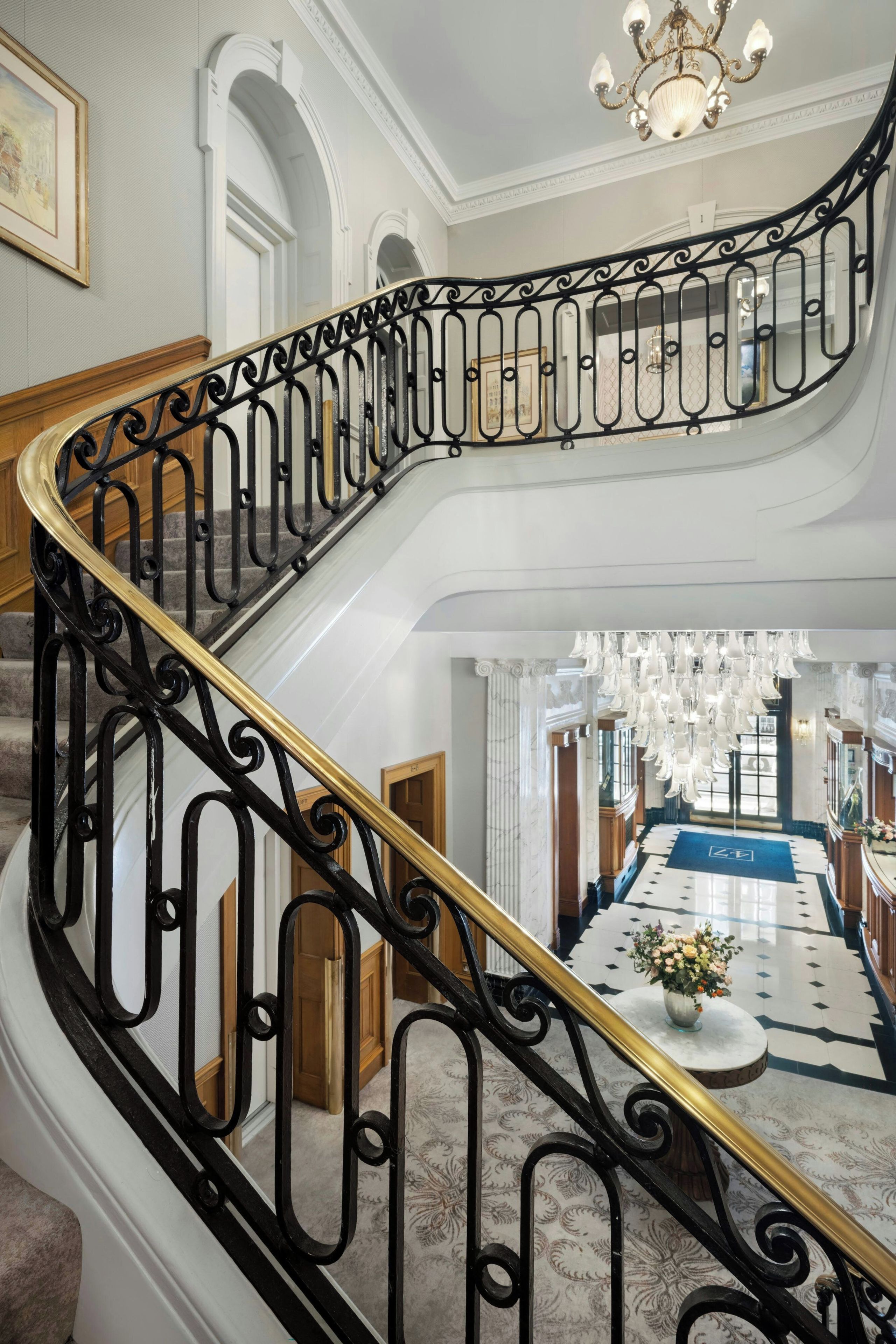 A grand staircase with ornate black metal railings and a golden handrail, leading to a well-lit upper floor with a chandelier.