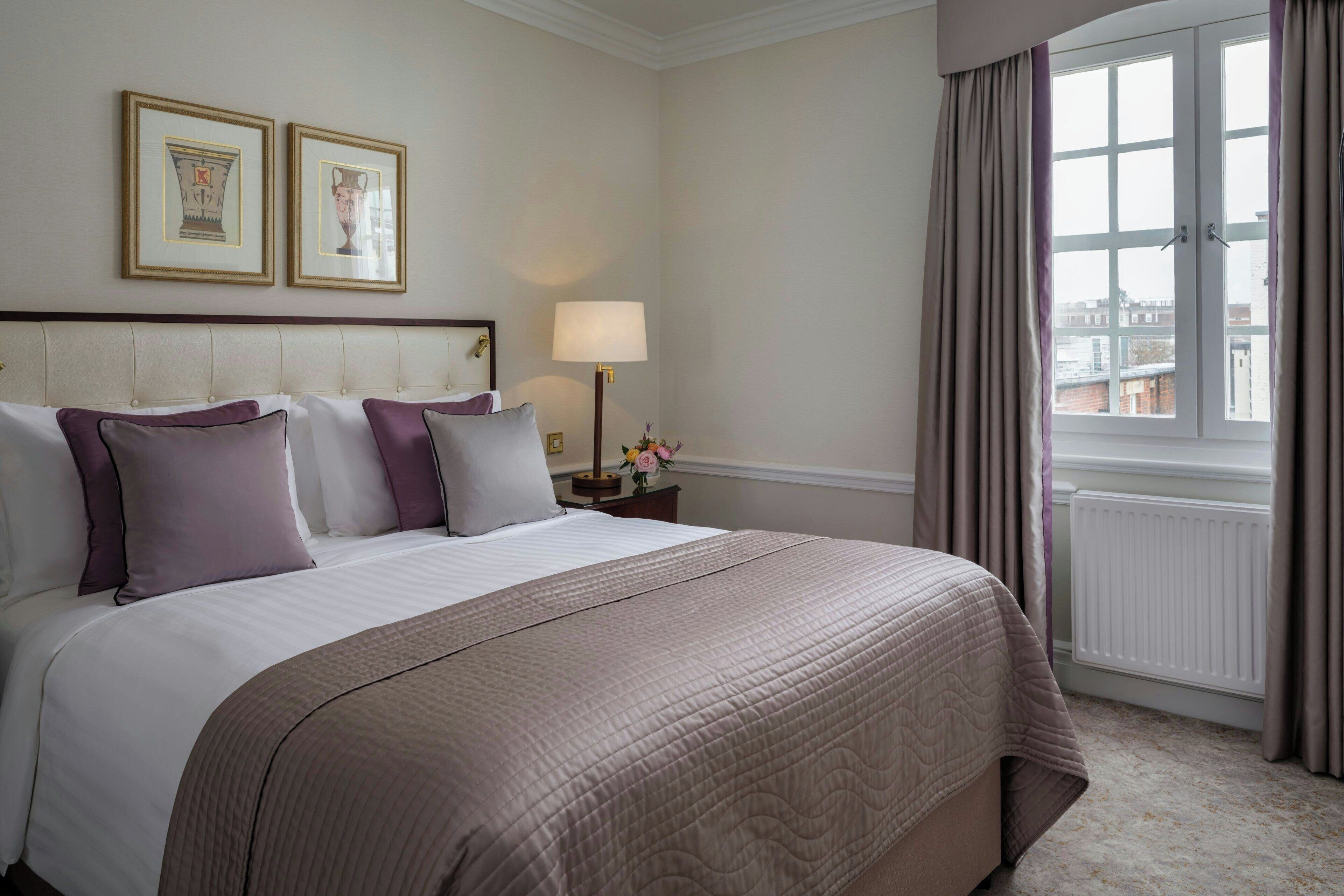 A hotel room with a large bed, a nightstand with a lamp, framed pictures on the wall, and a window with curtains.