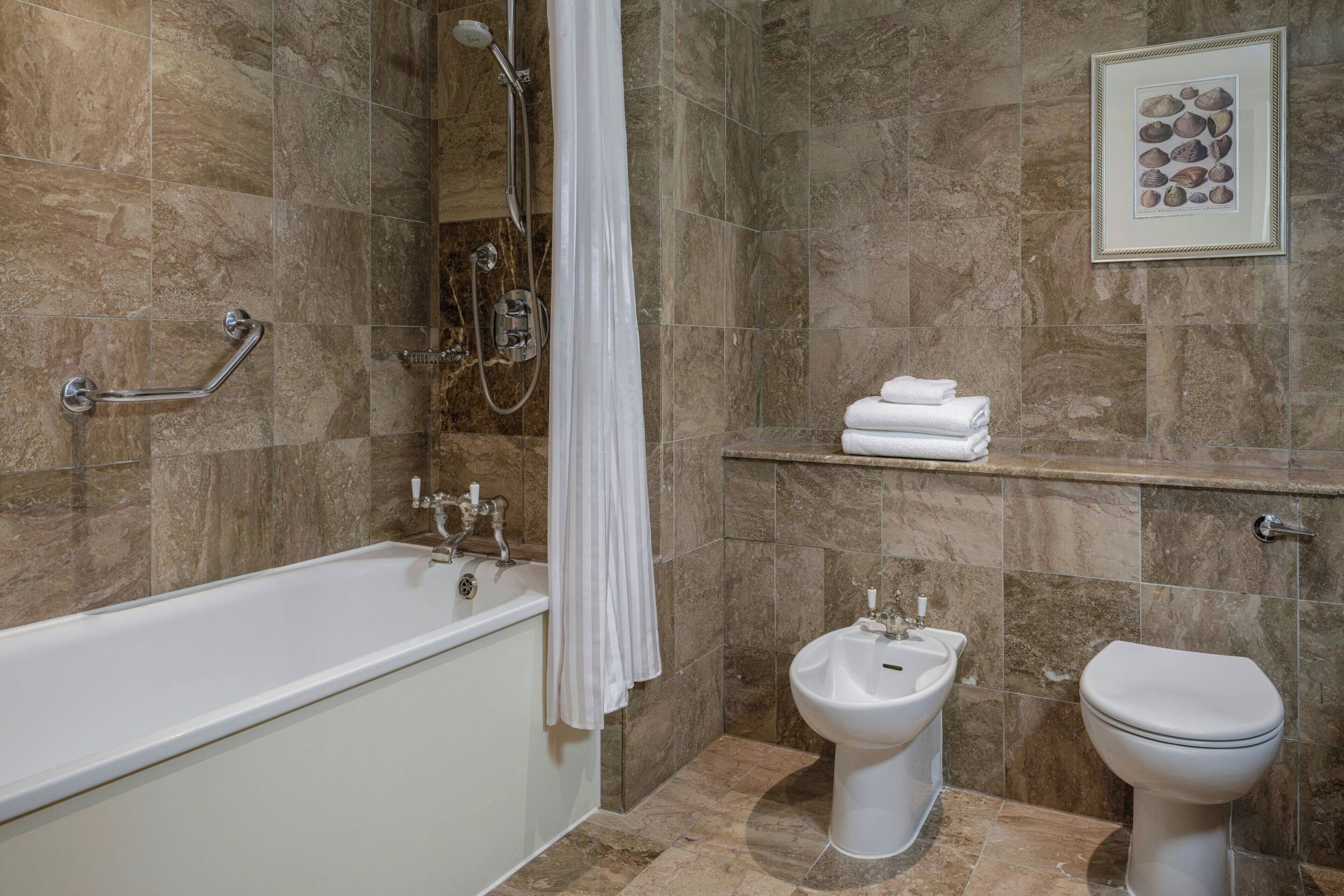 Baño con bañera, bidé e inodoro, todo en blanco, con cabezal de ducha y cortina a la vista.