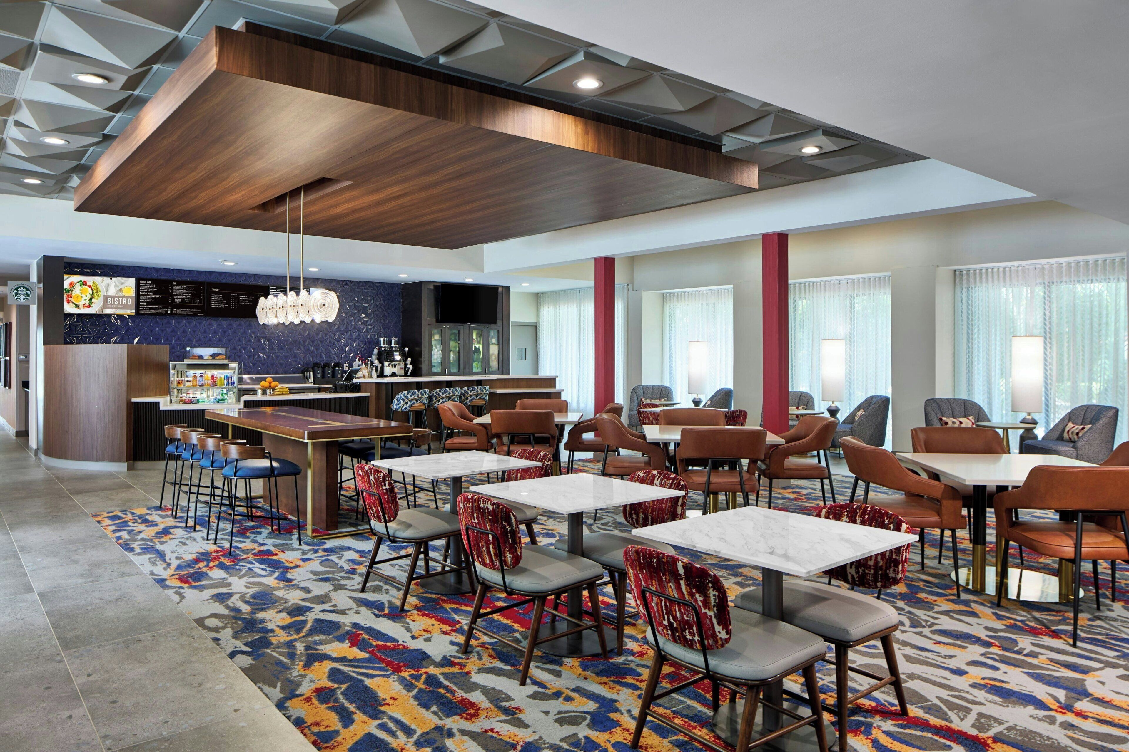 Vestíbulo de hotel con varias mesas de comedor, sillas, una zona de bar y una alfombra estampada.