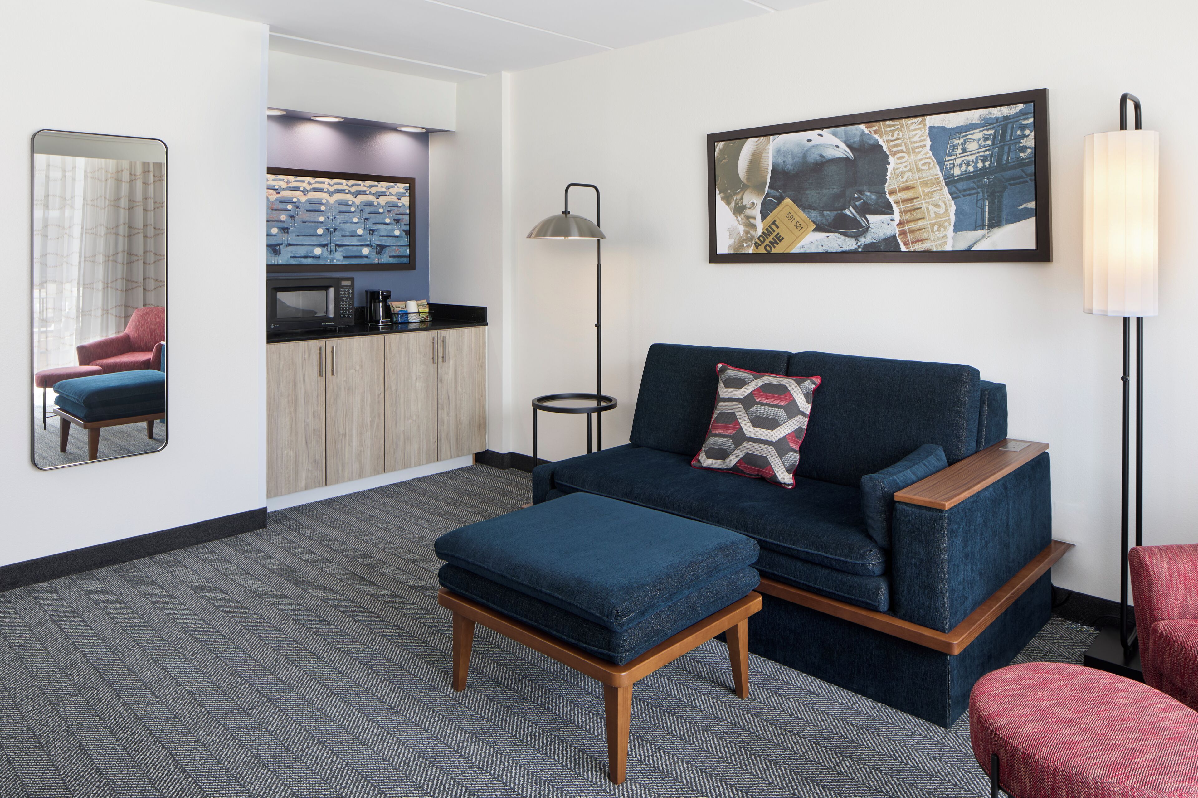 A modern hotel room with a sofa, ottoman, a small table, a coffee machine, a mirror, a painting, and a lamp.