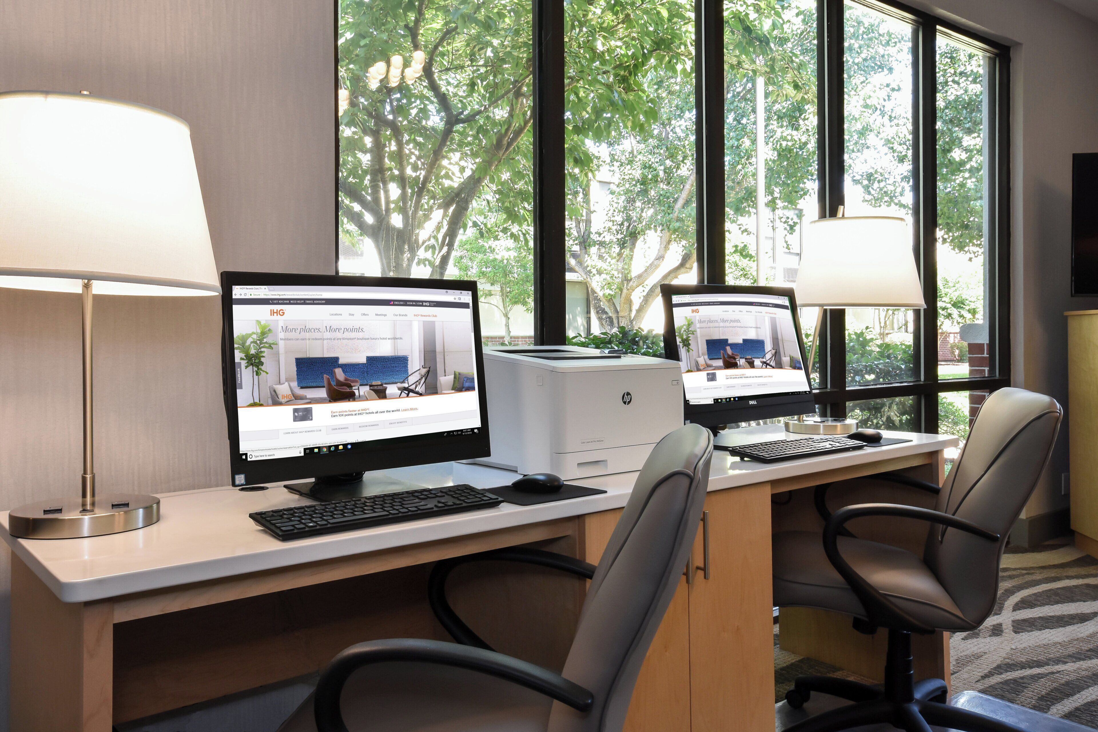 A hotel room with two desktop computers, a desk, chairs, and a window with a view of trees.