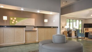 A modern hotel reception area with a reception desk, a seating area with a round ottoman, and a television. - Holiday Inn Lumberton by IHG (Lumberton)