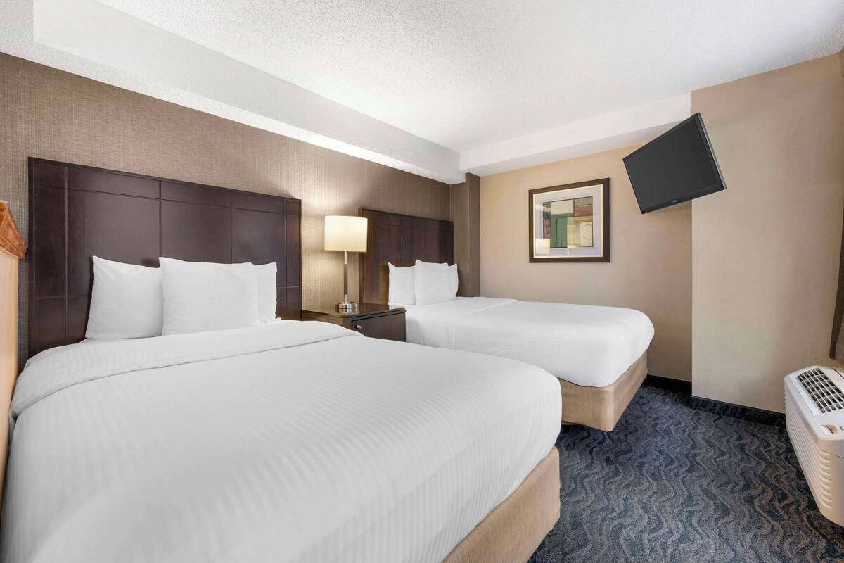 A hotel room with two beds, a flat-screen TV mounted on the wall, a bedside table with a lamp, and a portable heater.