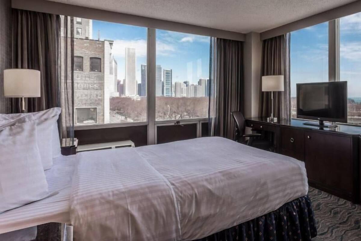A hotel room with a large bed, a desk, a chair, a TV, and a view of a cityscape.