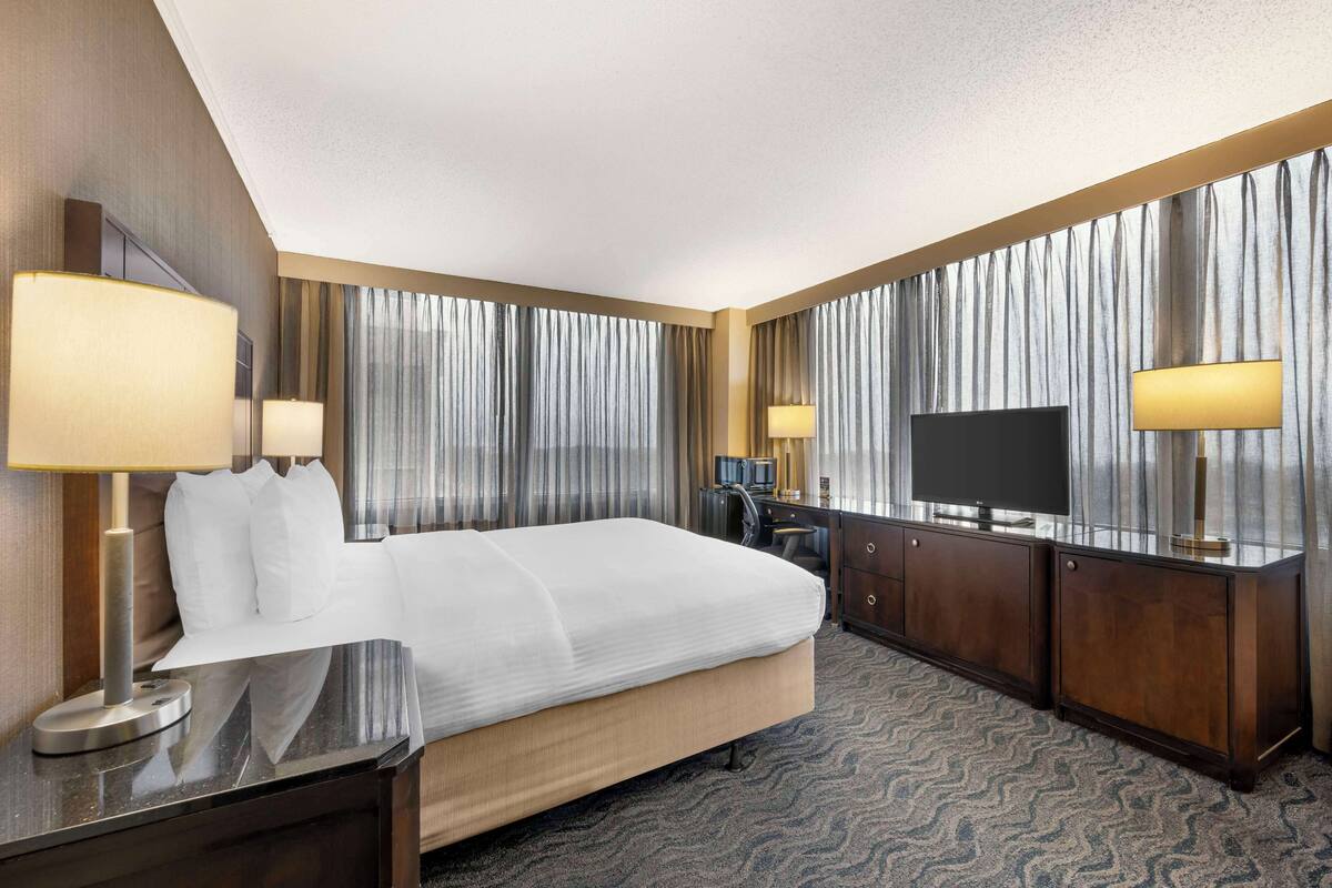 A hotel room with a large bed, two bedside tables with lamps, a TV, a desk with a chair, and a wardrobe.