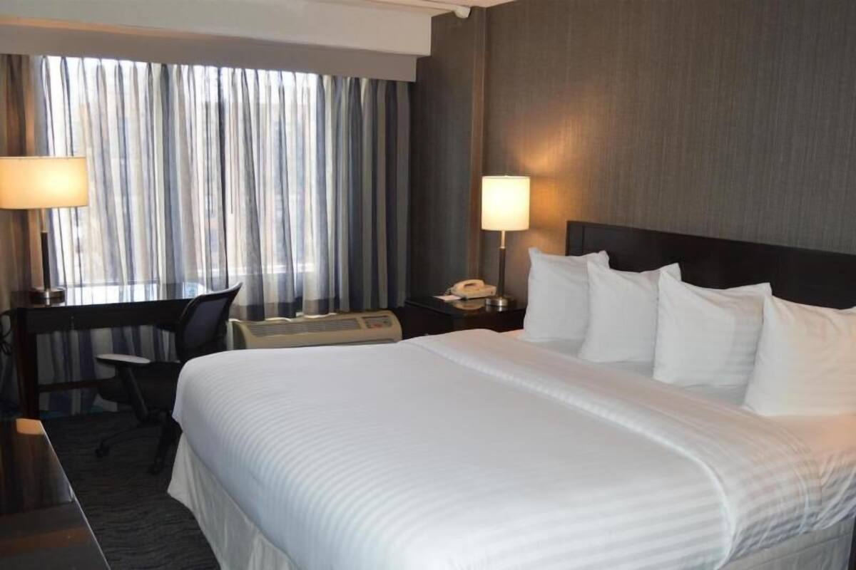 A hotel room with a large bed, a desk, a chair, a desk lamp, a phone, and a wall-mounted heater.
