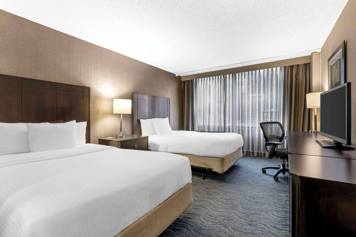 A hotel room with two beds, a desk, a TV, and a large window with curtains.