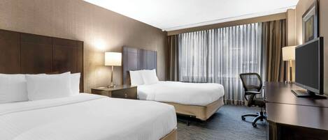 A hotel room with two beds, a desk, a TV, and a large window with curtains.