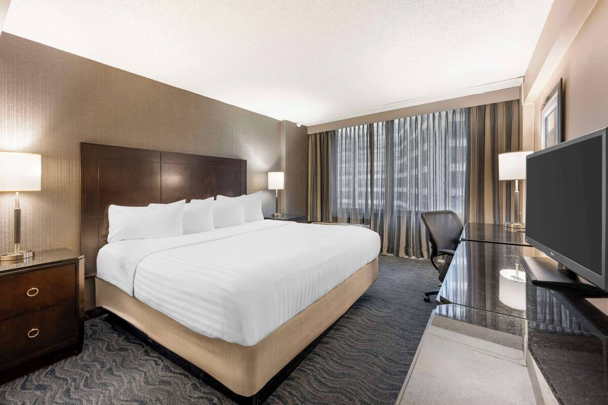 A hotel room with a large bed, a desk with a flat-screen TV, a chair, and a window with curtains.