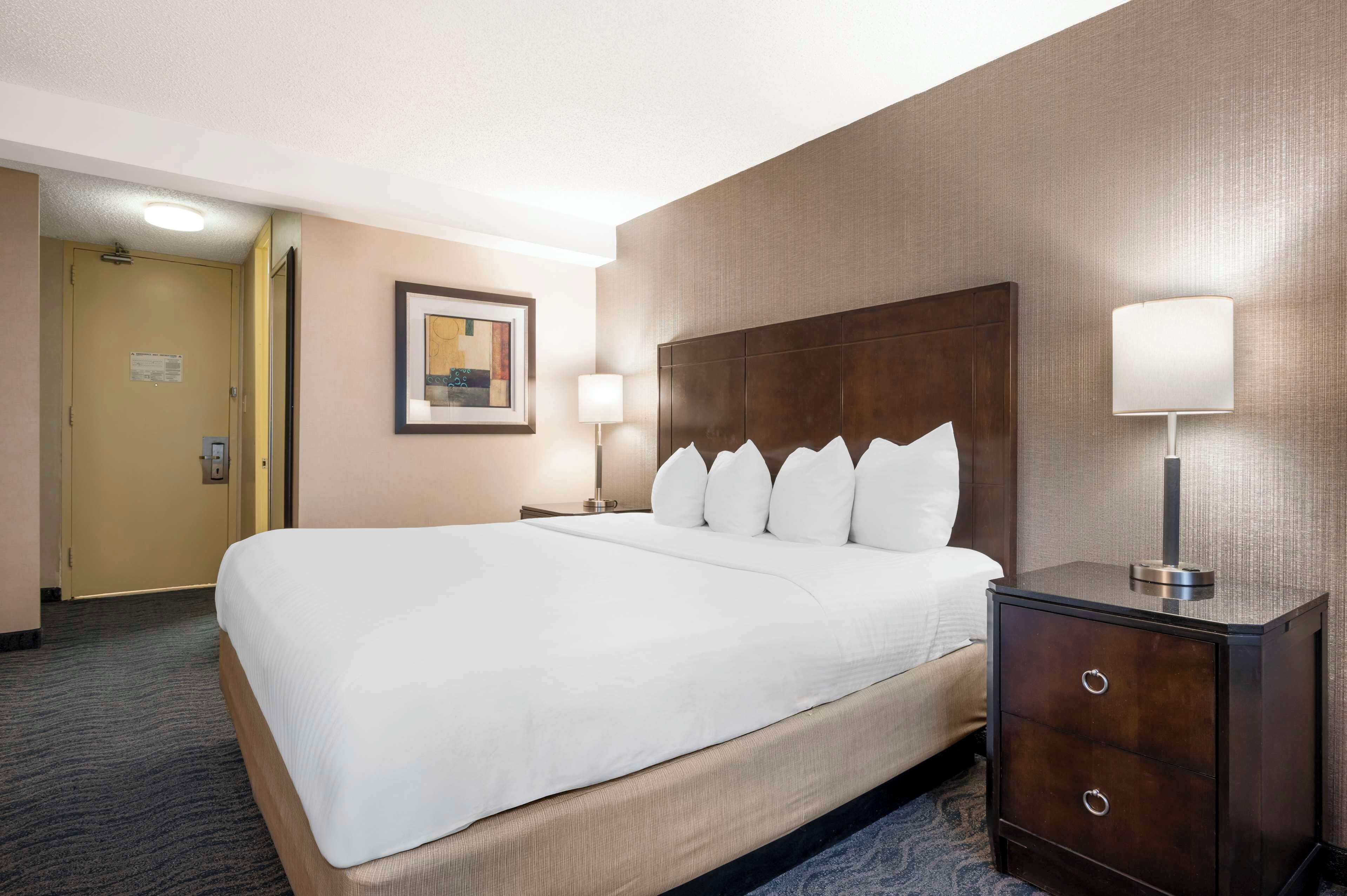 Quarto de hotel com uma cama grande, duas mesas de cabeceira com abajures, uma porta e um quadro na parede.