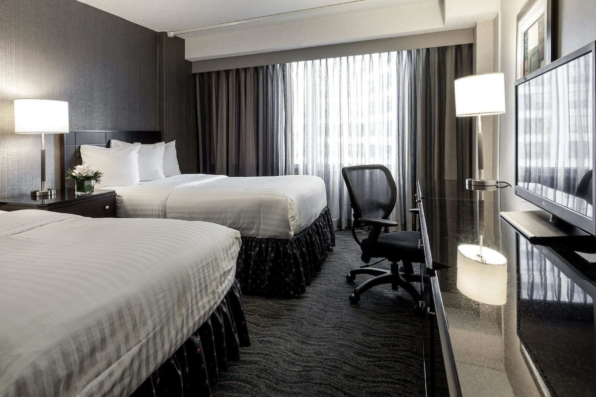 Een hotelkamer met twee bedden, een bureau met stoel, een televisie en een groot raam met gordijnen.