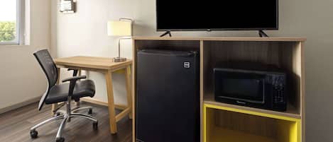 A compact hotel room with a wooden desk, a black office chair, a flat-screen TV mounted on the wall, a black refrigerator, and a microwave on a yellow shelf.
