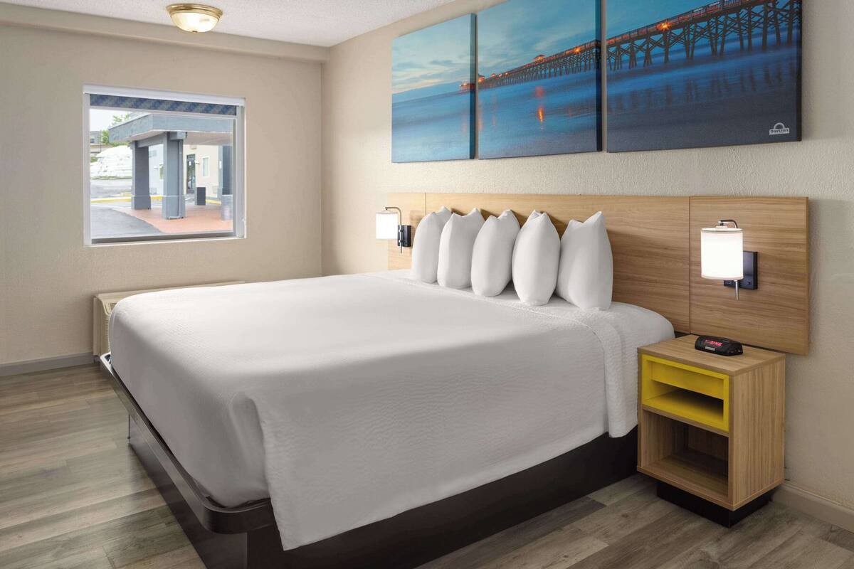 Habitación de hotel con una cama grande, una mesita de noche con lámpara y ventana con vista a un edificio.