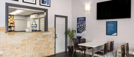 Un vestíbulo de hotel con mostrador de piedra, una recepción, un área de comedor con mesa y sillas, y un televisor montado en la pared.