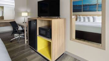 Een hotelkamer met een bed, een televisie op een houten standaard, een bureau met een stoel, een magnetron en een geel opbergmeubel.
