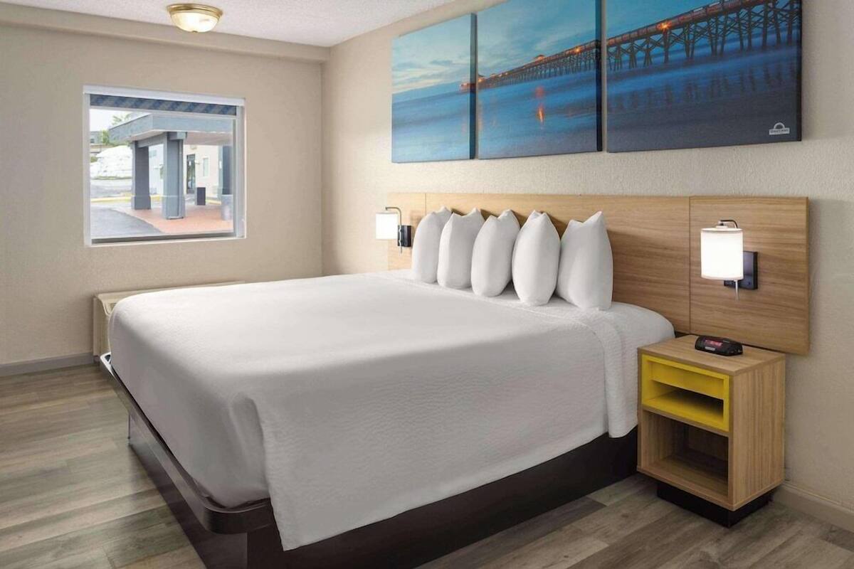 Habitación de hotel con cama, mesita de noche con lámpara, ventana con vistas y una obra de arte en la pared.