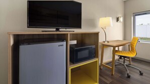 Habitació d'hotel senzilla amb televisió de pantalla plana, microones, nevera, una cadira groga, escriptori i làmpada.