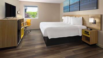 A modern hotel room with a large bed, a desk with a computer, a chair, and a window with a view of greenery.