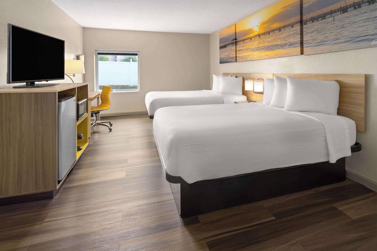 Habitación de hotel con dos camas, un escritorio con televisor, una silla y un cuadro de un atardecer sobre un muelle.